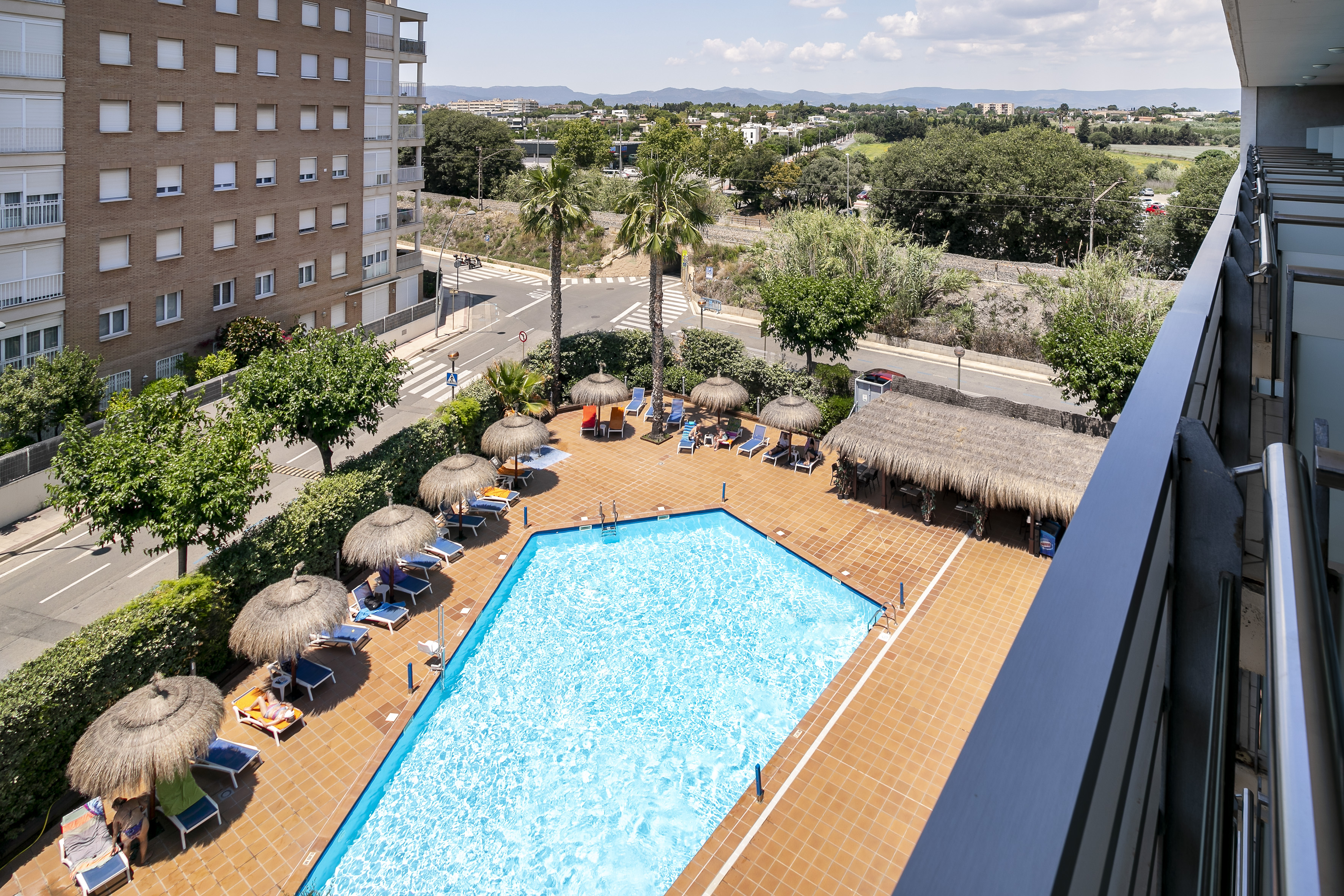Hotel outdoor swimming pool with straw umbrellas, sun loungers, and distant city.