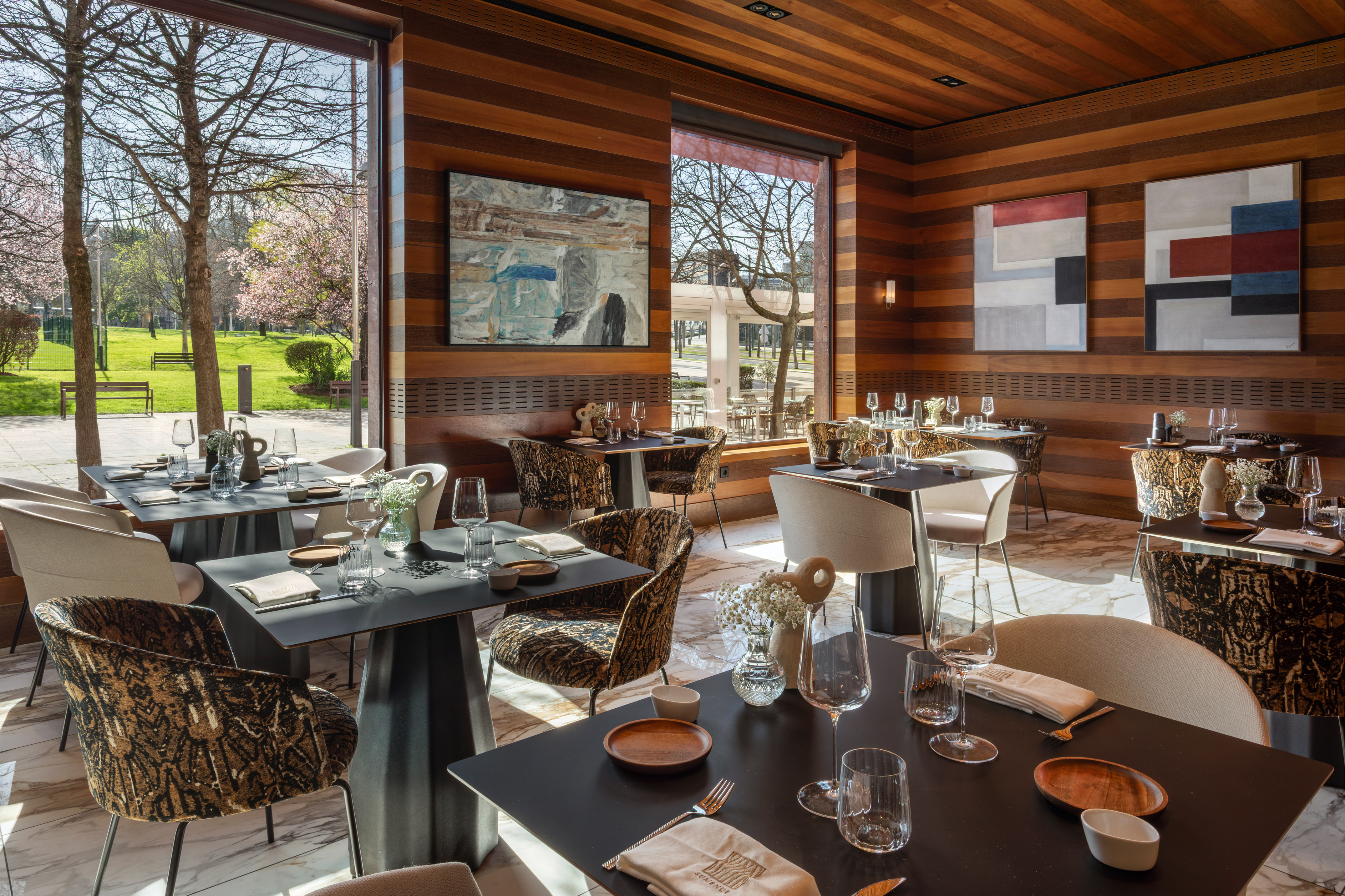 Modern luxury restaurant dining room with park view, wooden interior, and set tables.