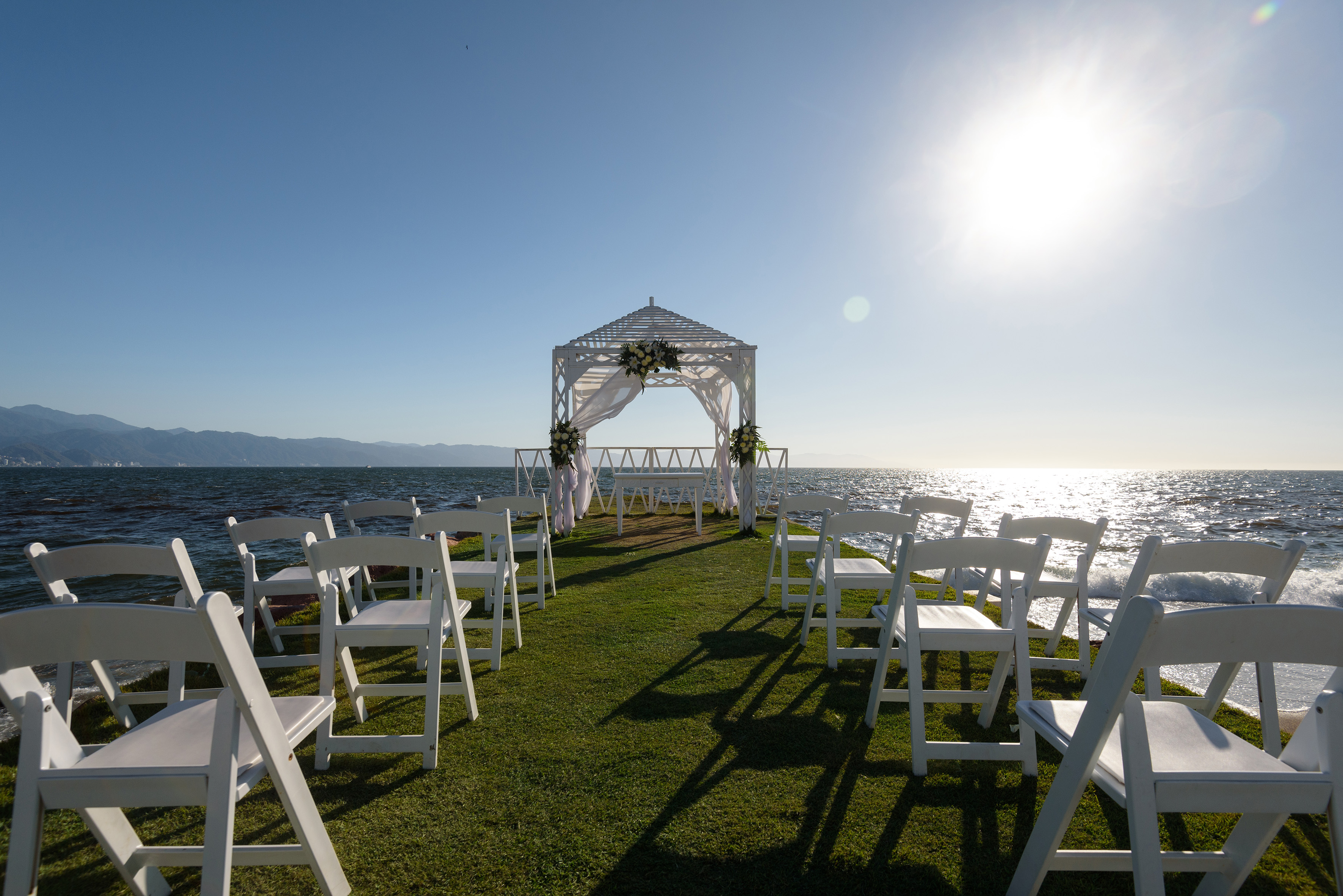 Oceanfront wedding ceremony setup with white chairs and floral gazebo.