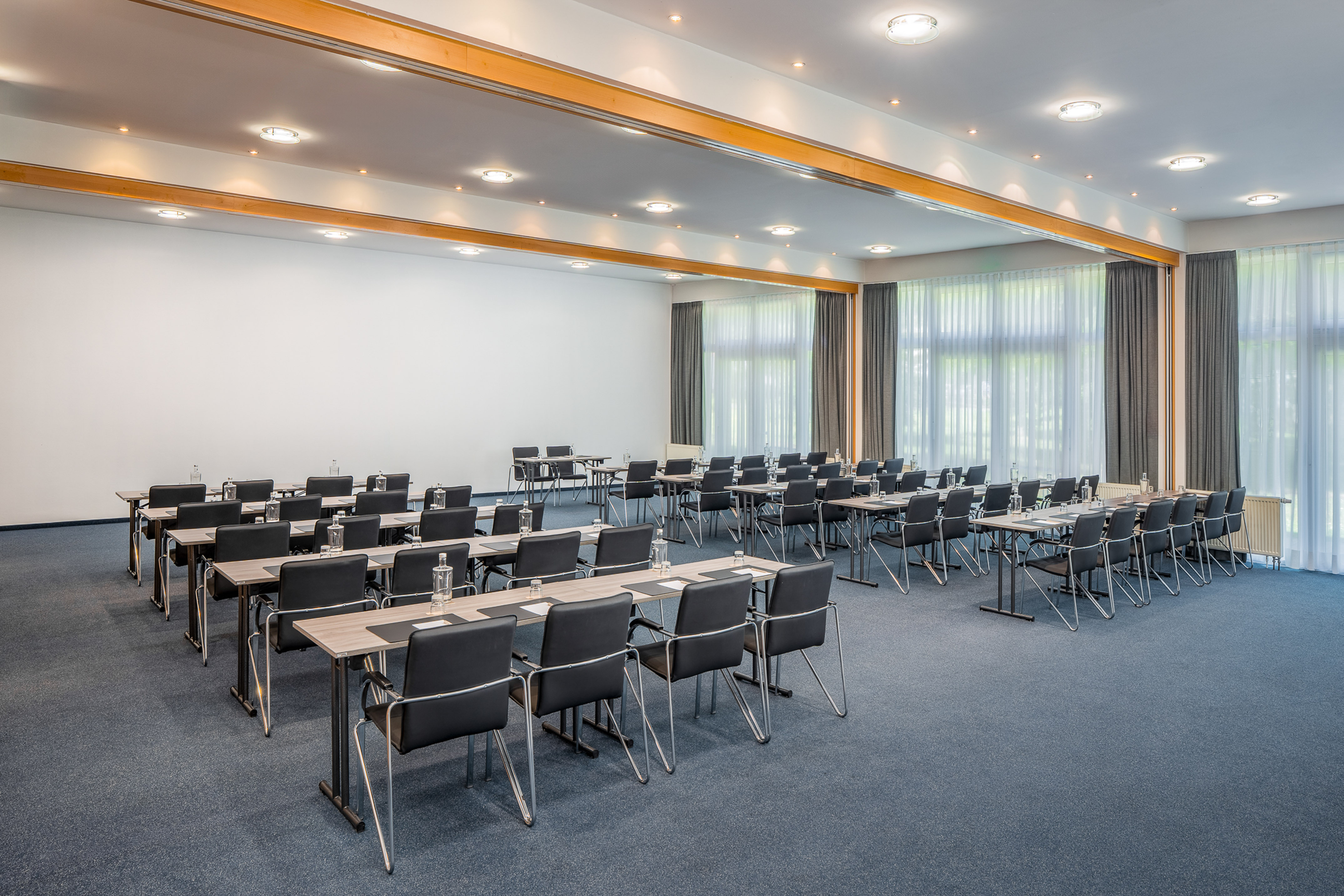 Modern conference room with tables, chairs, water bottles. Blue carpet, bright windows.