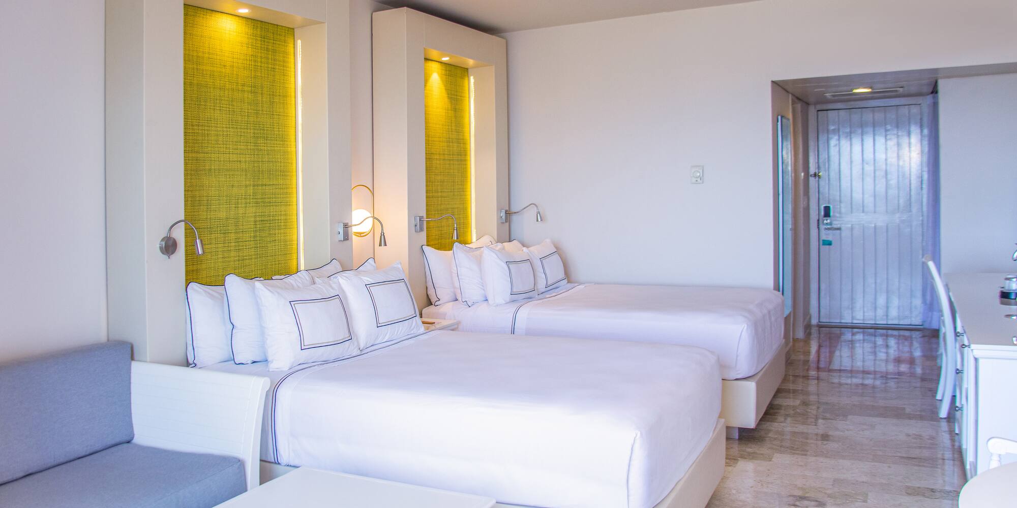 Modern hotel room with two beds, white linens, green accents, and a sofa.