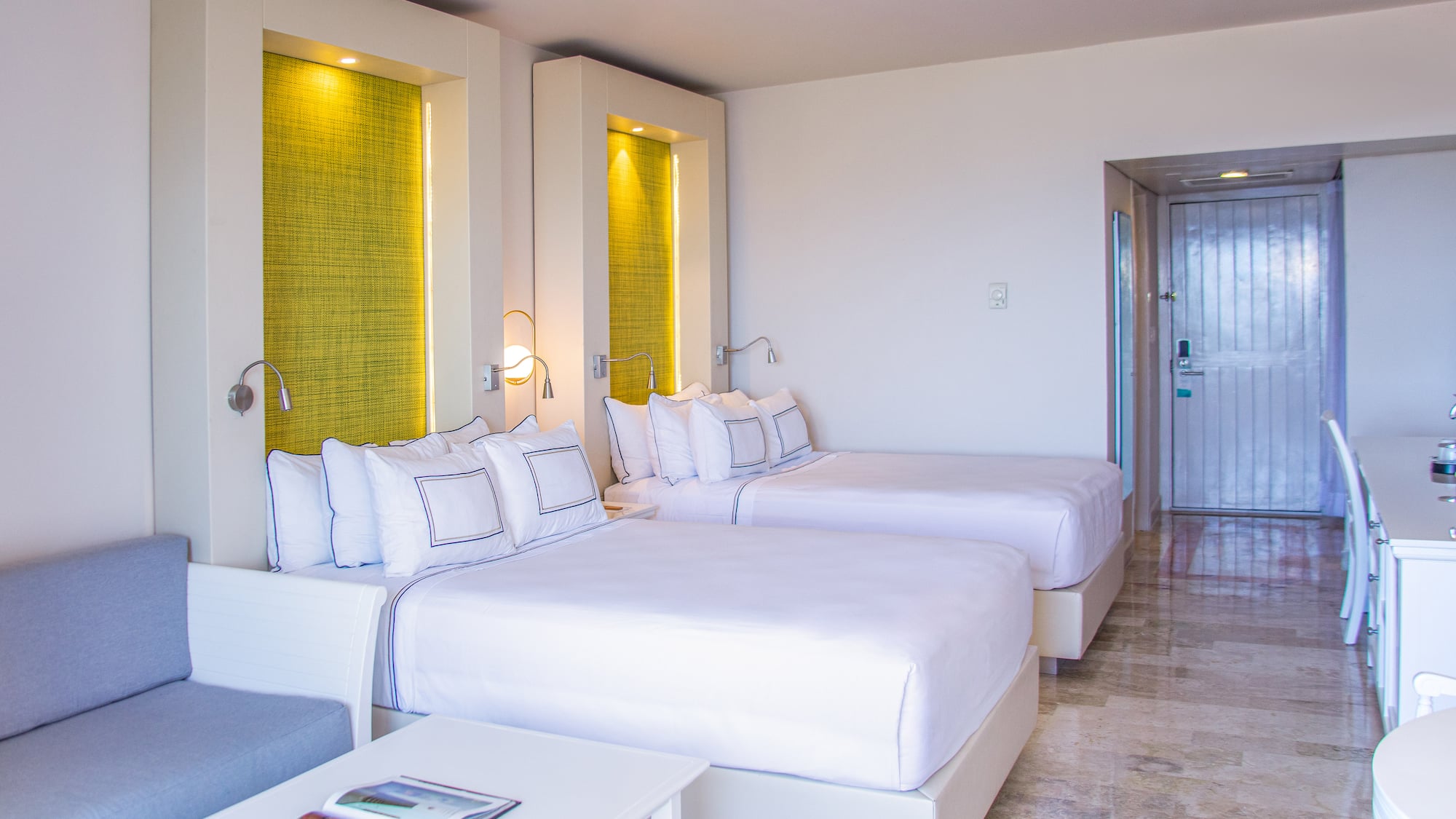 Bright hotel twin room with white beds, green lit headboards, and a sofa.