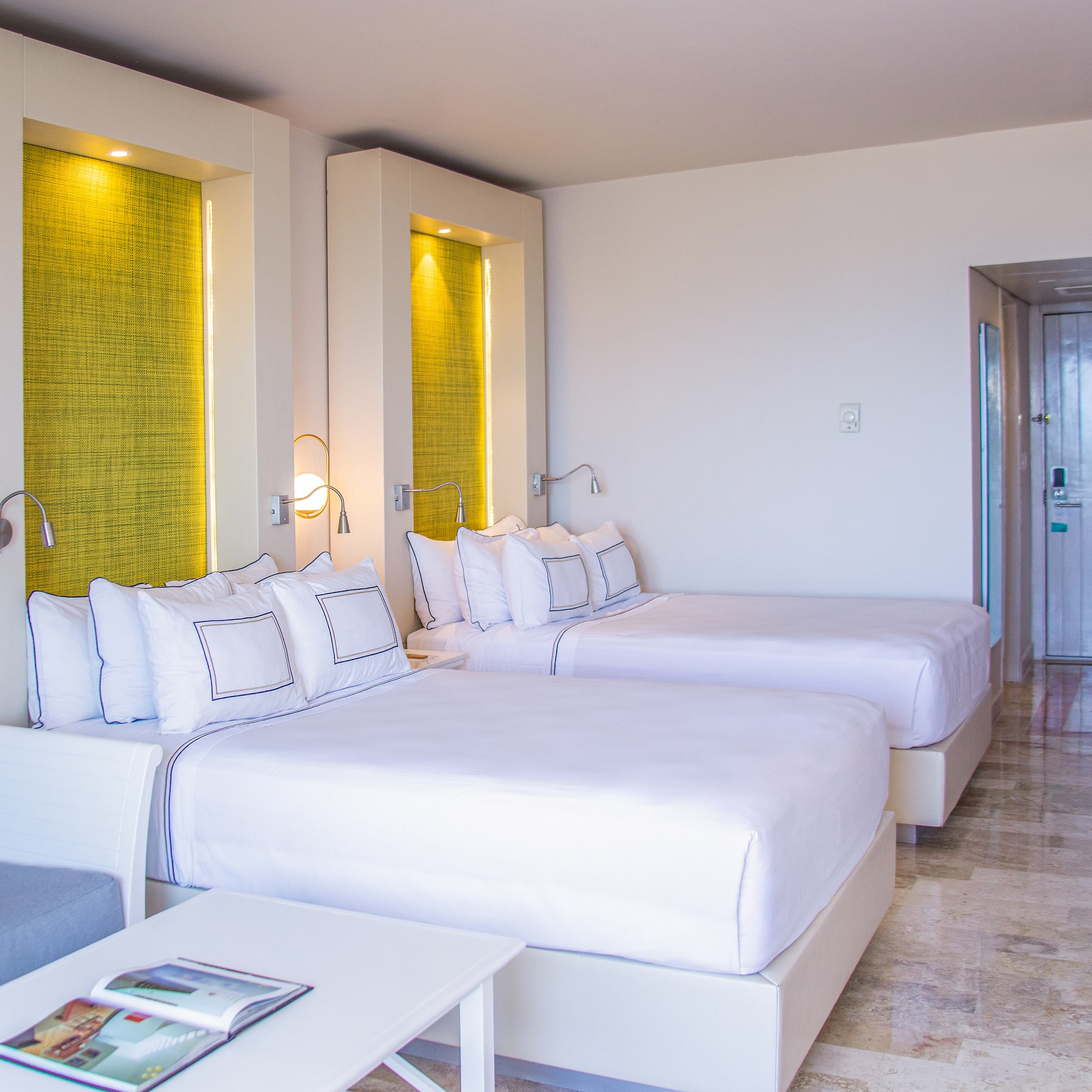 Modern hotel room with two beds, white linens, green accents, and a sofa.