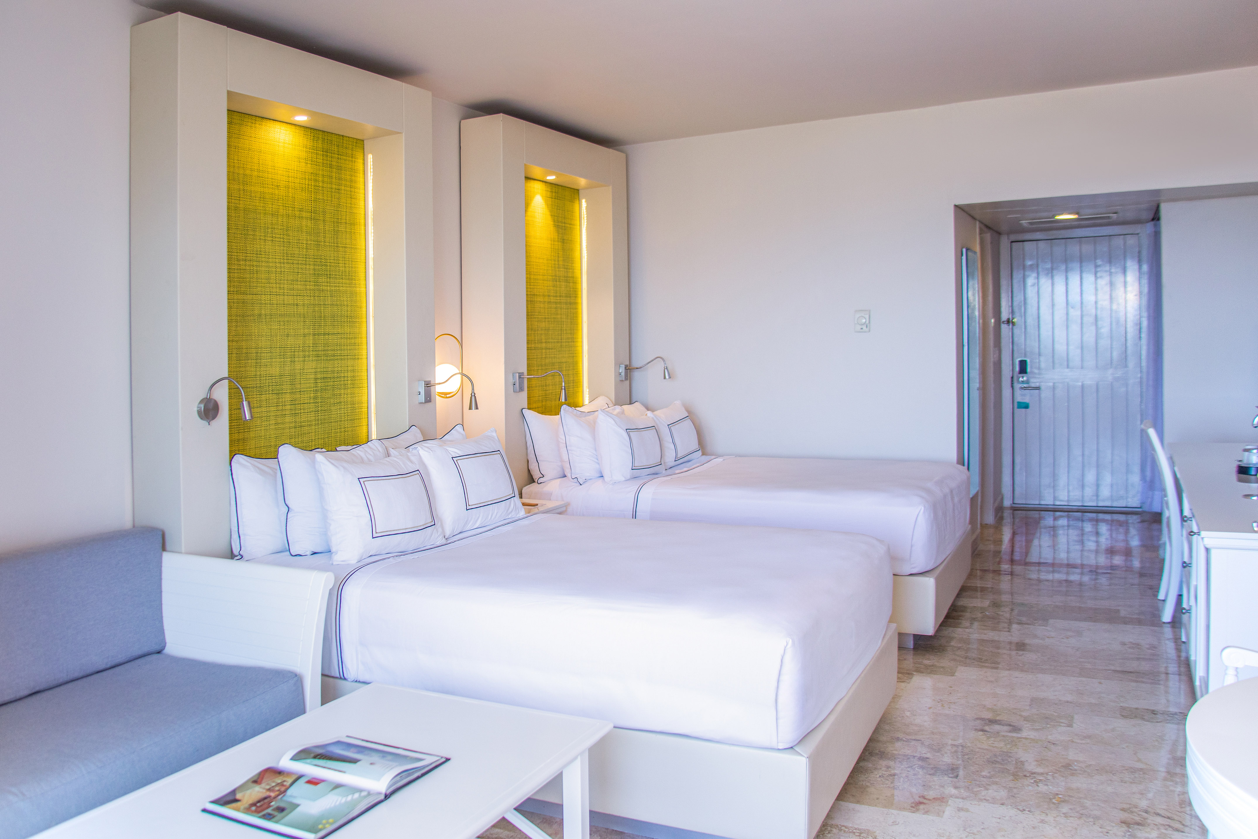 Modern hotel room with two beds, white linens, green accents, and a sofa.