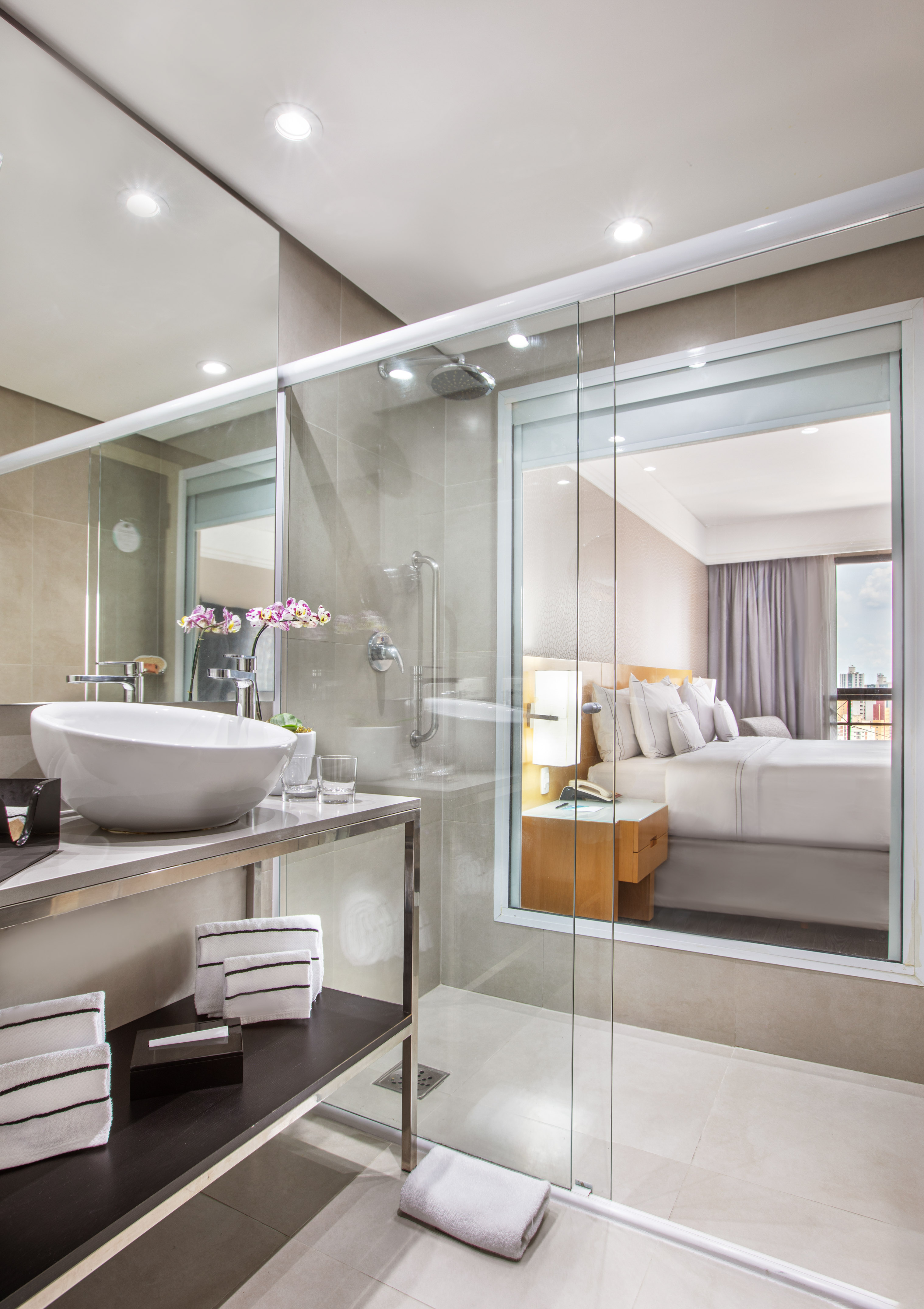 Modern bathroom with glass shower and vanity overlooking a hotel bedroom.