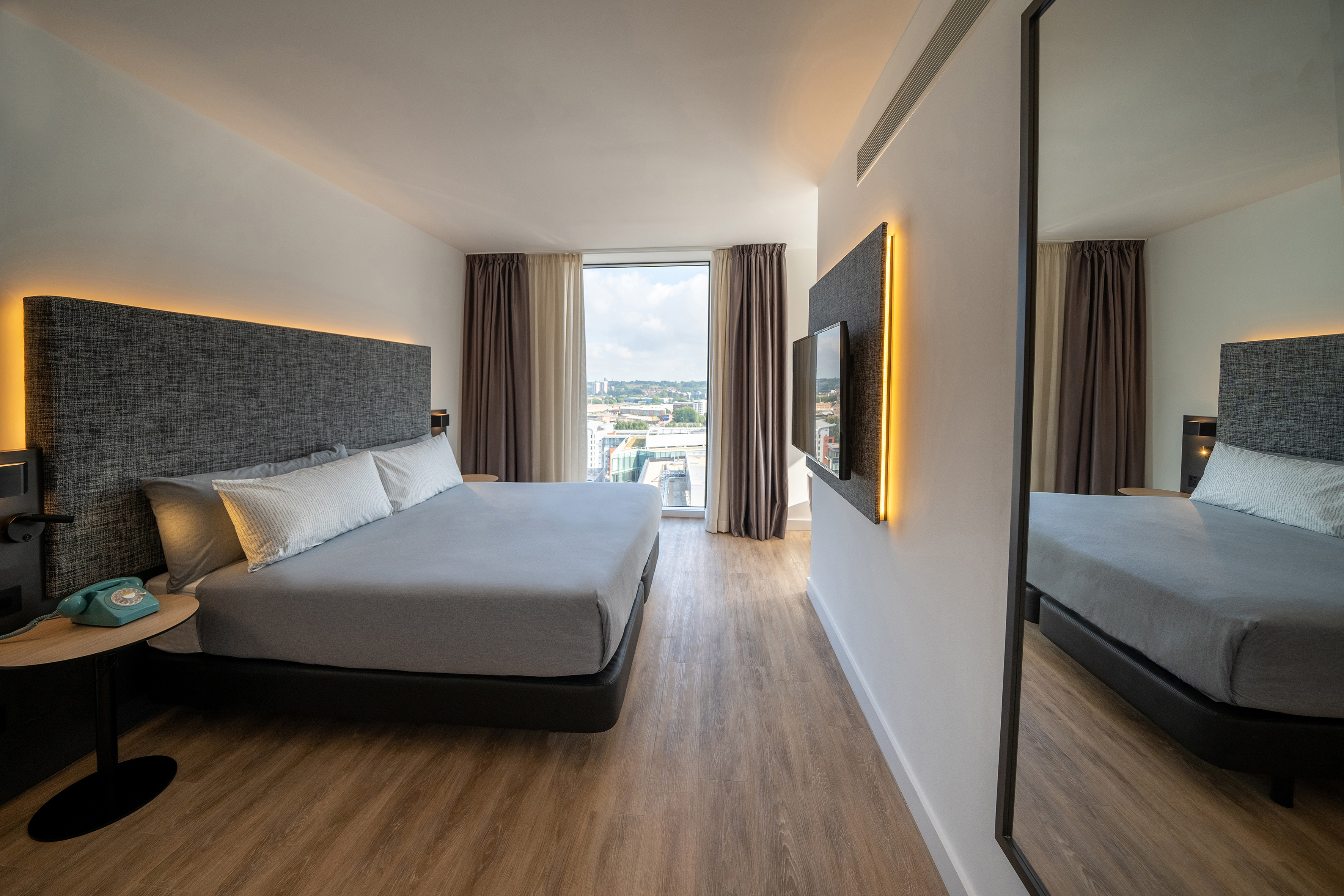 Modern hotel room with king bed, city view window, wood floor, and ambient lighting.