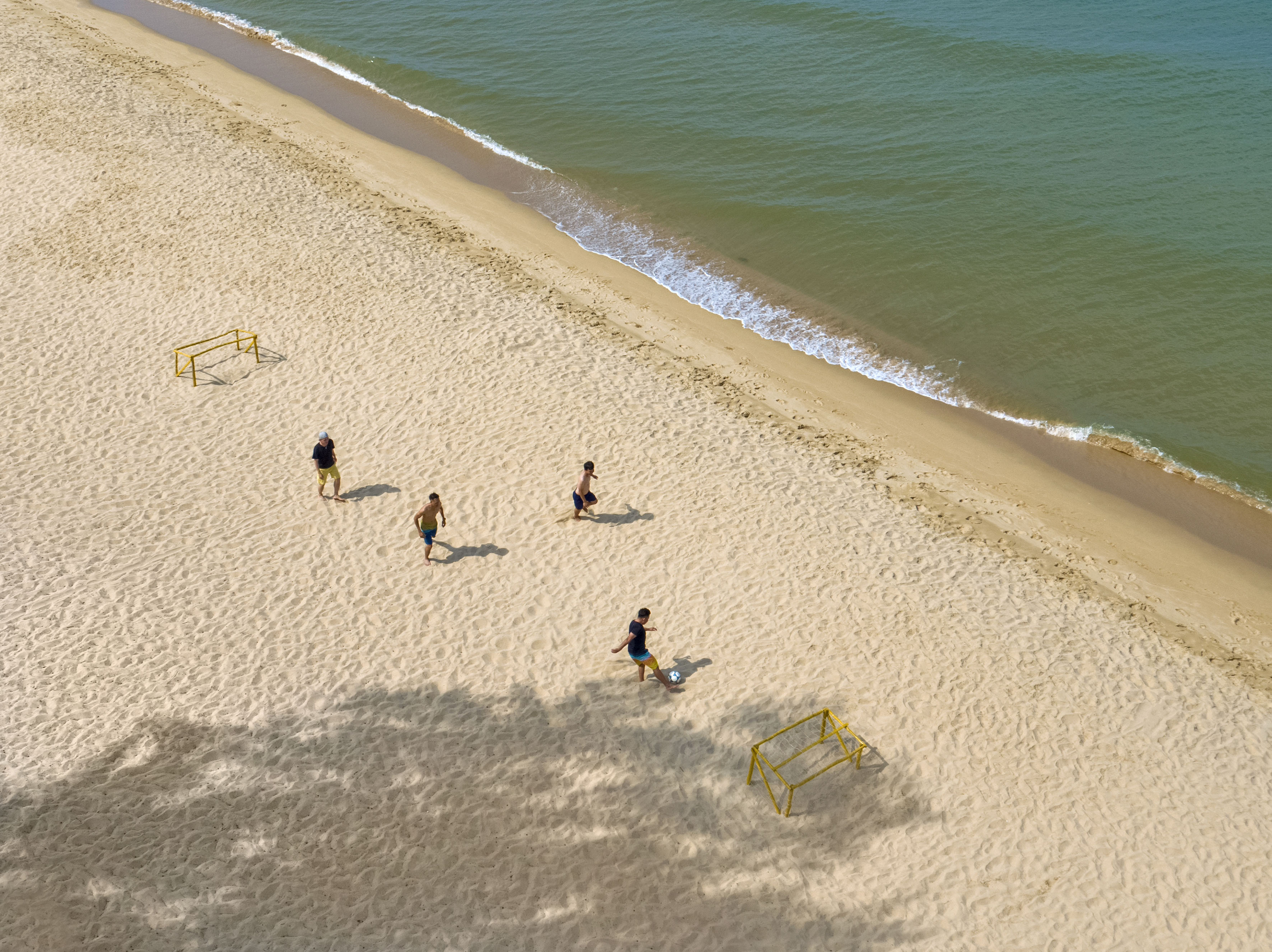 Beach soccer game with four men on sand beside ocean, two yellow goals.