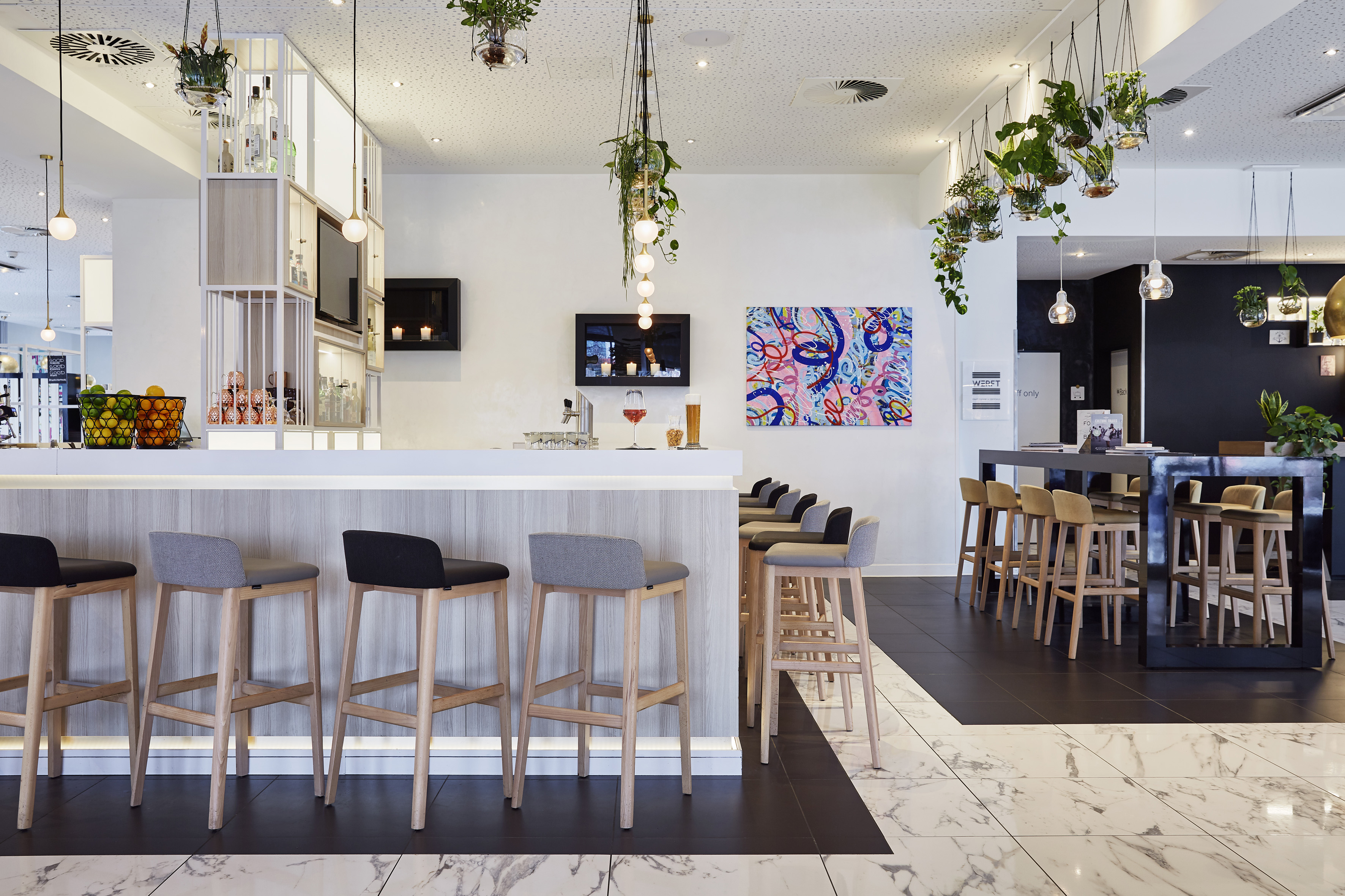 Modern hotel bar with white counter, wooden stools, hanging plants, and abstract art.