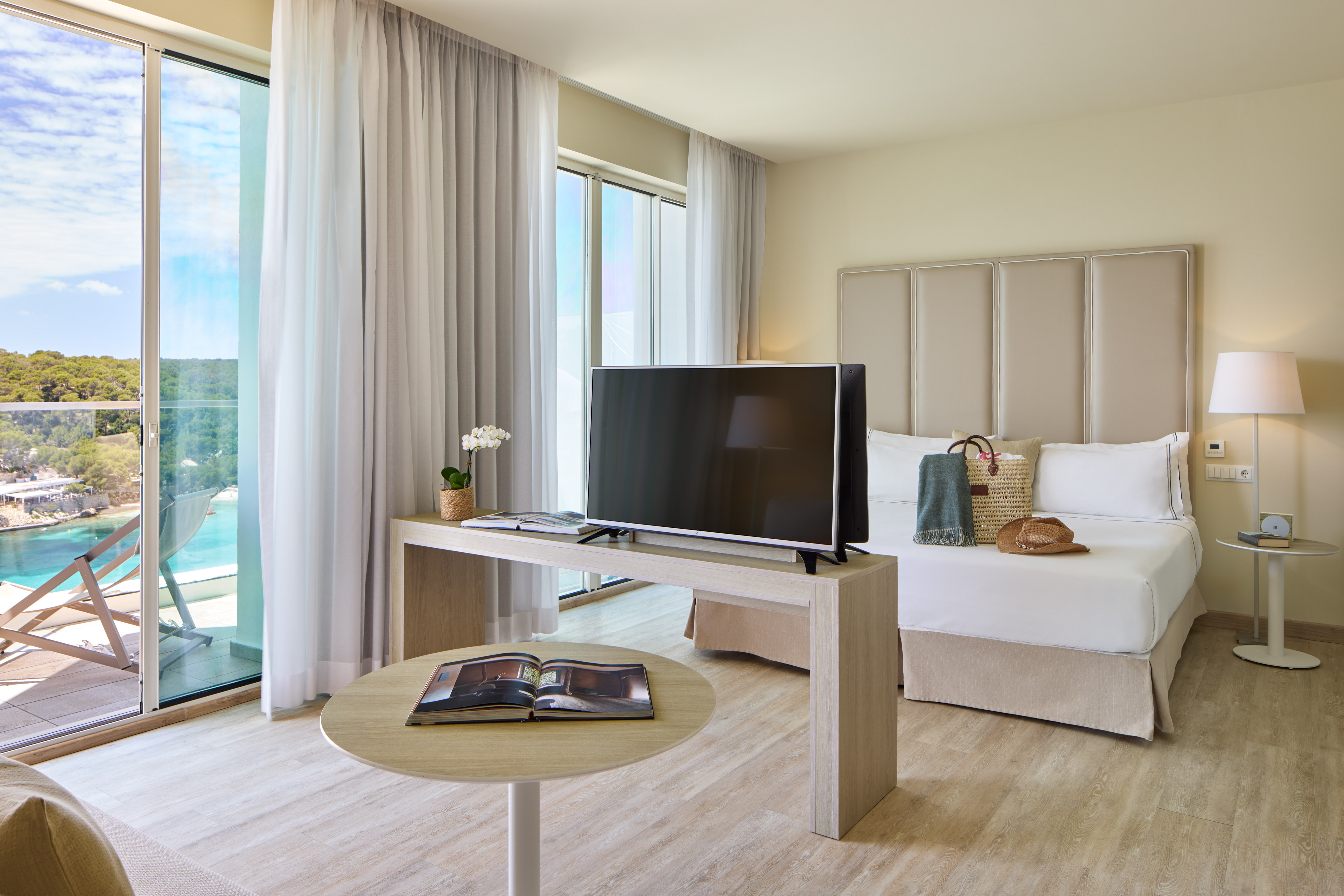 Modern hotel room with balcony overlooking a bay, large bed, and TV.