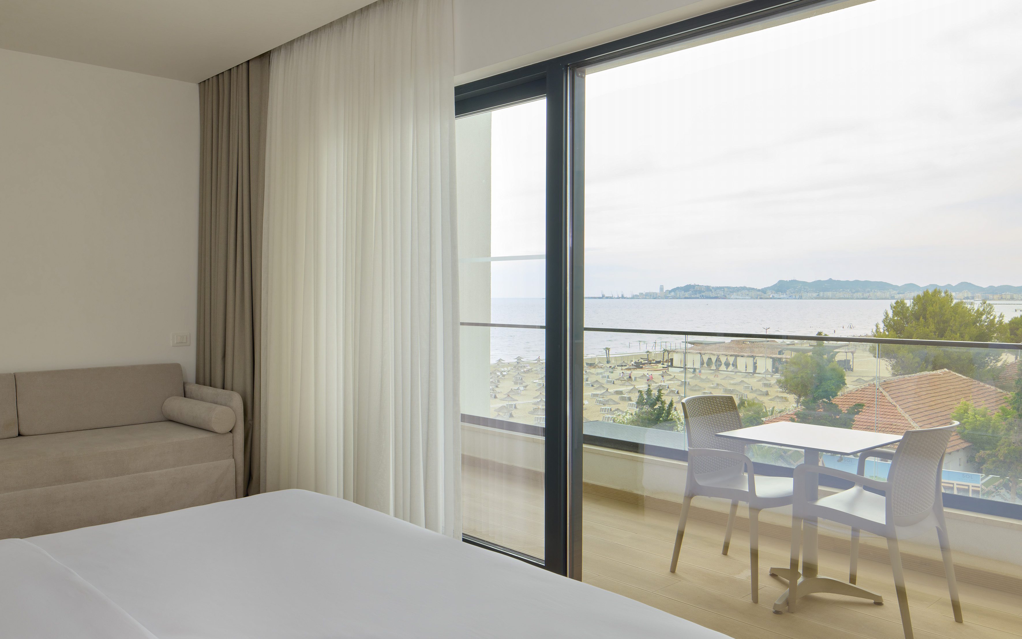 Hotel room with bed, sofa, and balcony with beach and sea view.
