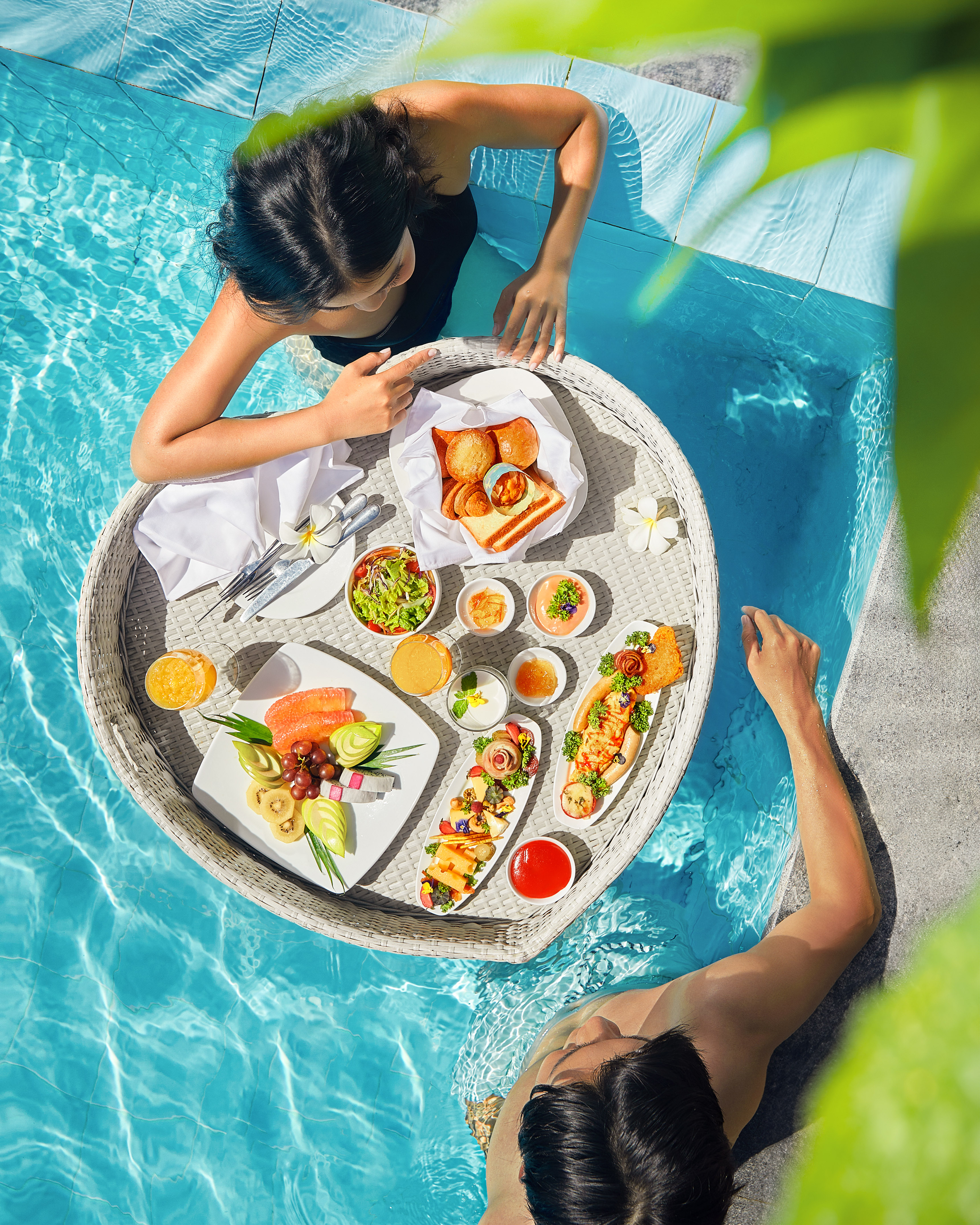 Couple enjoying a luxury floating breakfast spread in a sunny blue pool.
