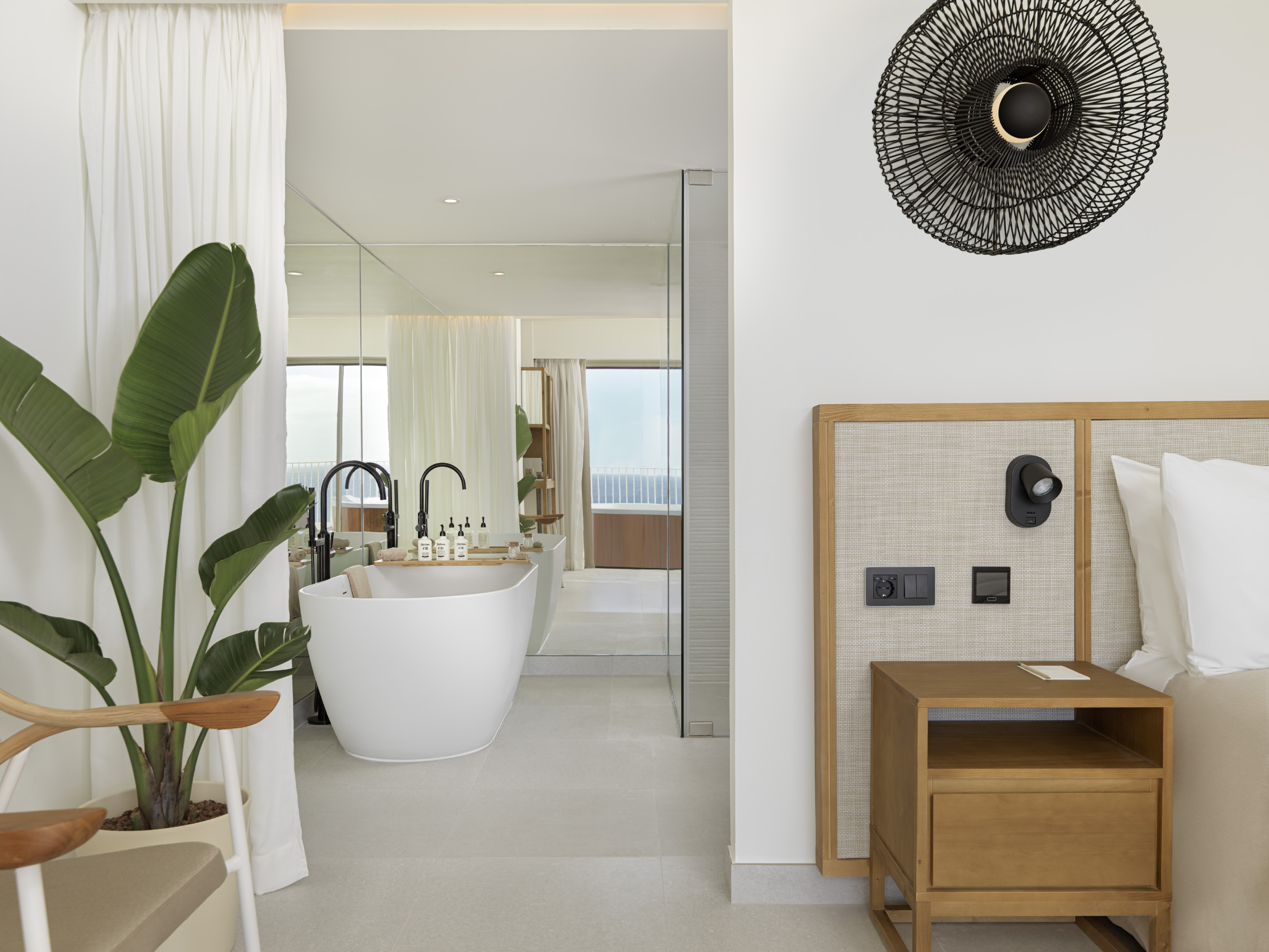 Hotel suite: modern bedroom, open bathroom, freestanding tub, potted plant.