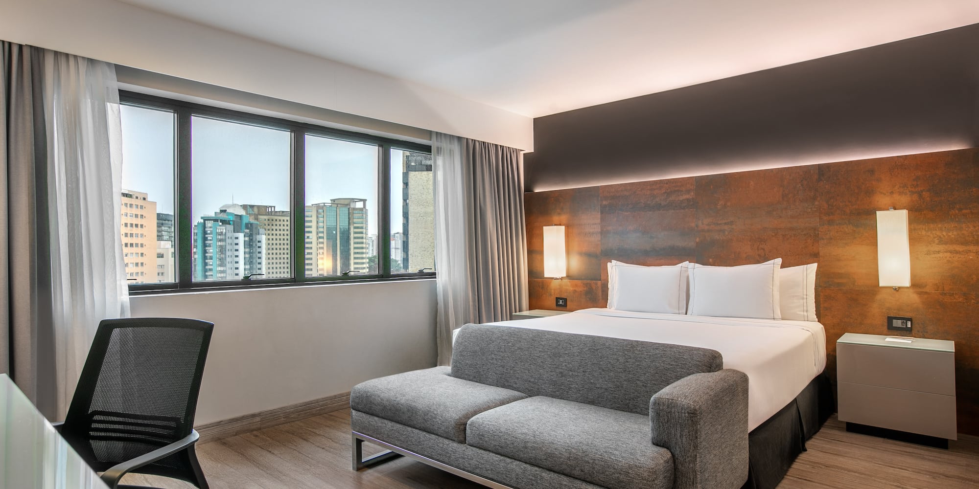 Quarto de hotel moderno com cama de casal, sofá cinza, escrivaninha e janela com vista para a cidade.