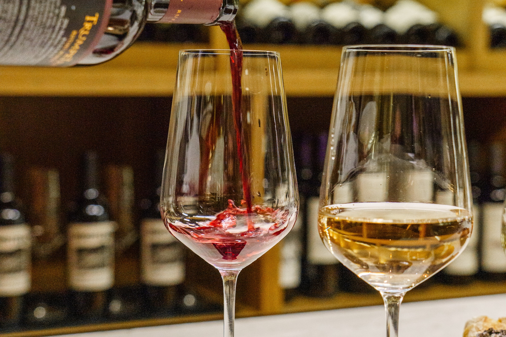 Red wine pouring into a glass, golden wine in another. Wine bottles on shelves.