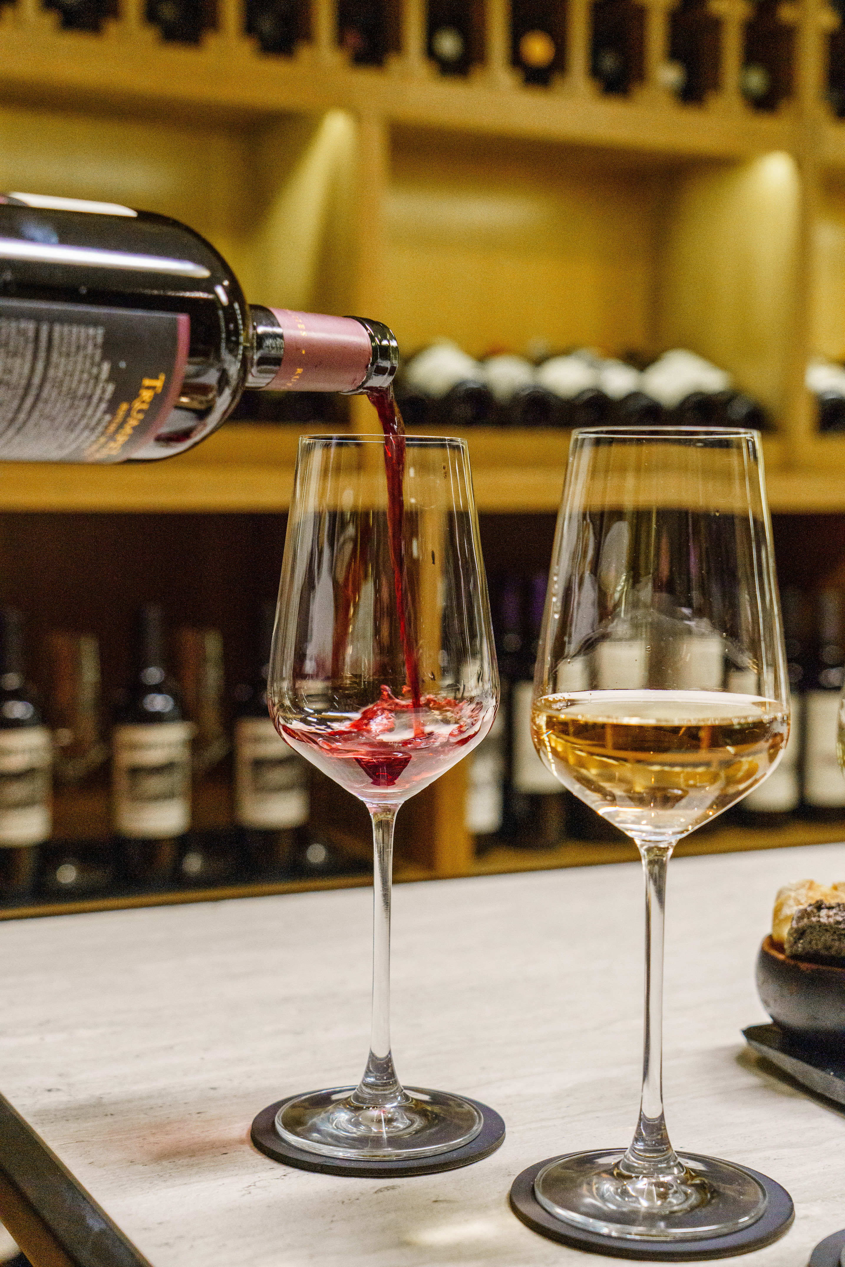 Red wine pouring into a glass, golden wine in another. Wine bottles on shelves.