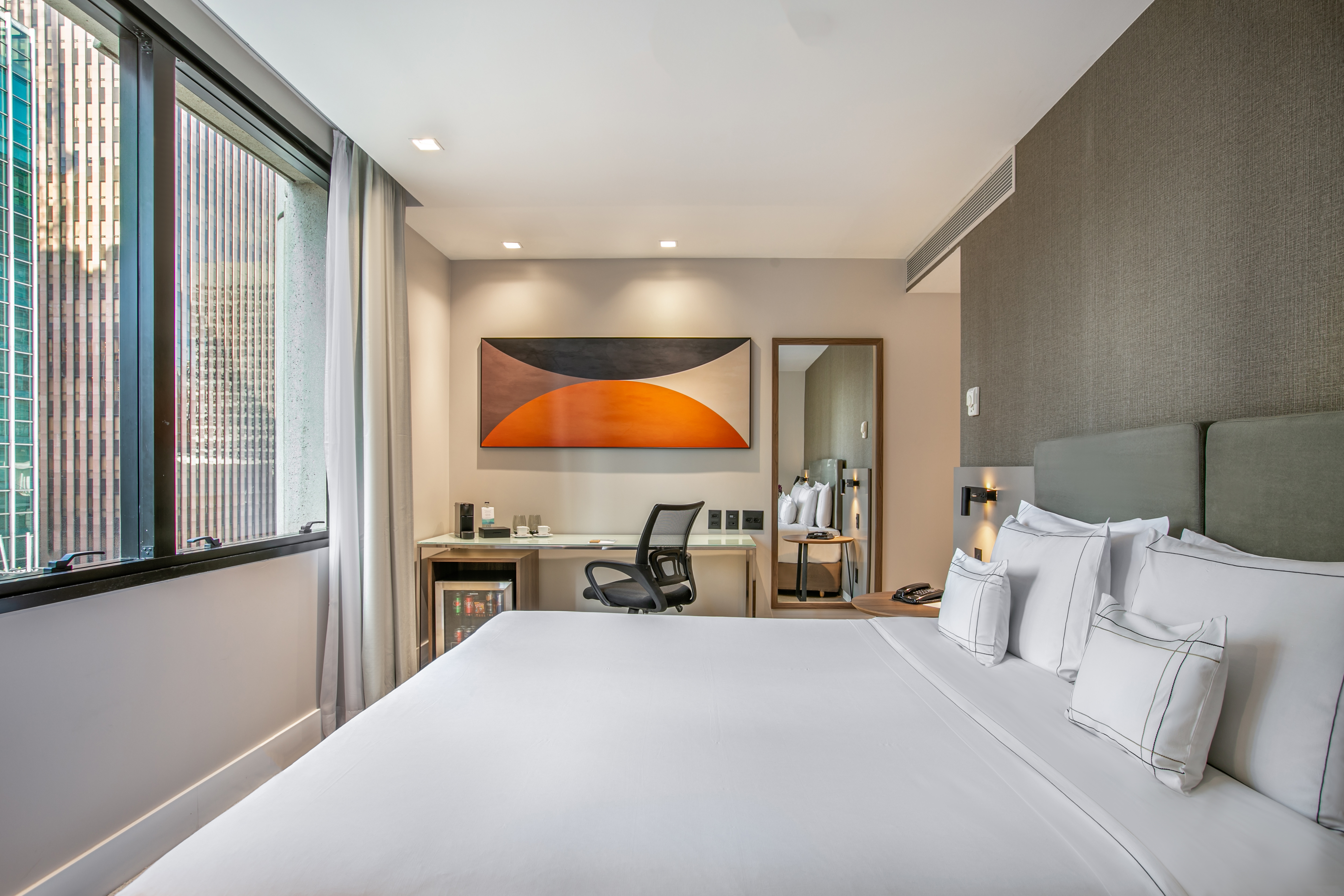 Modern hotel room with large bed, desk, minibar, and city view window.