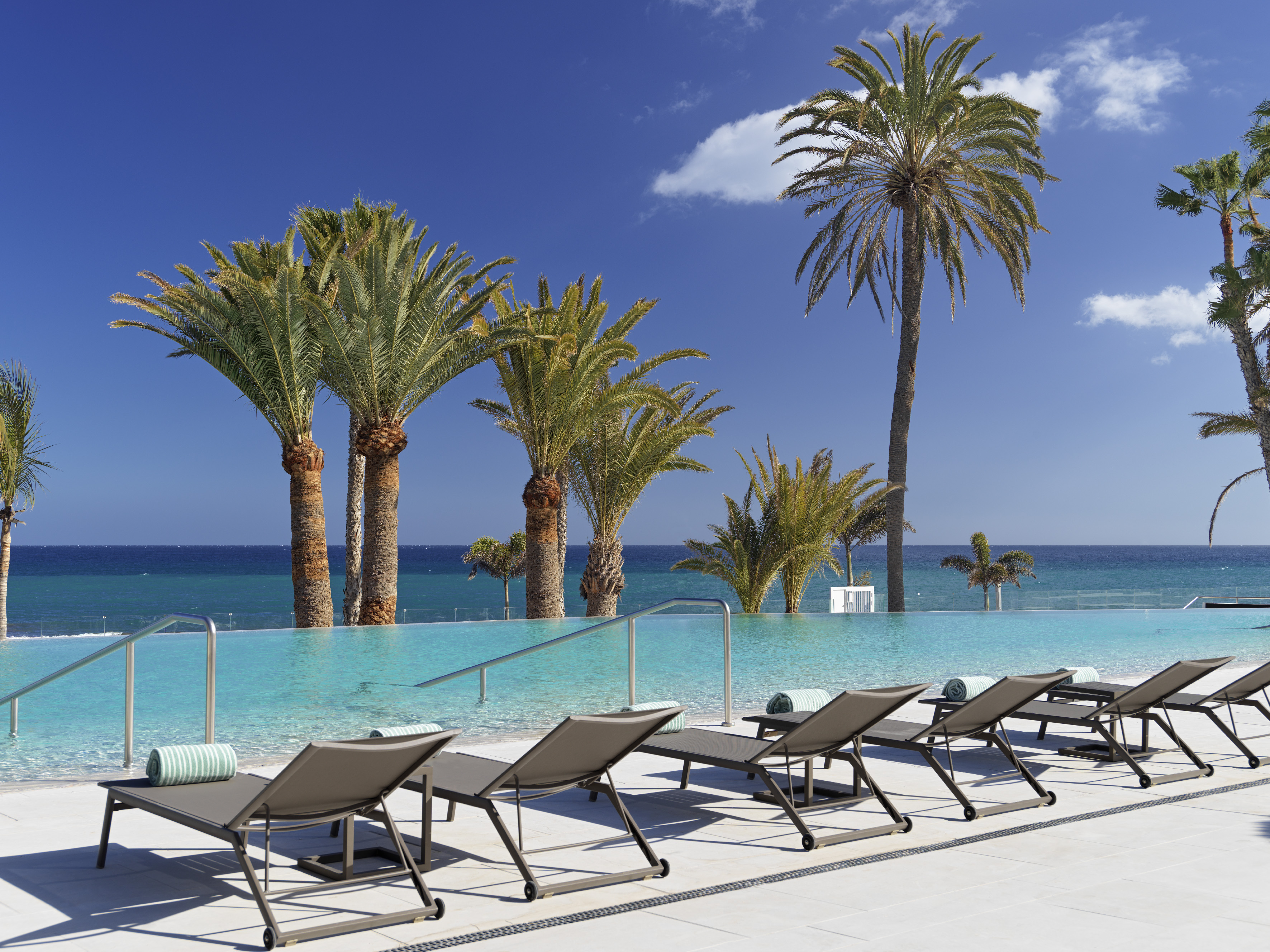 Resort infinity pool with ocean view, tropical palm trees, and sunbeds.