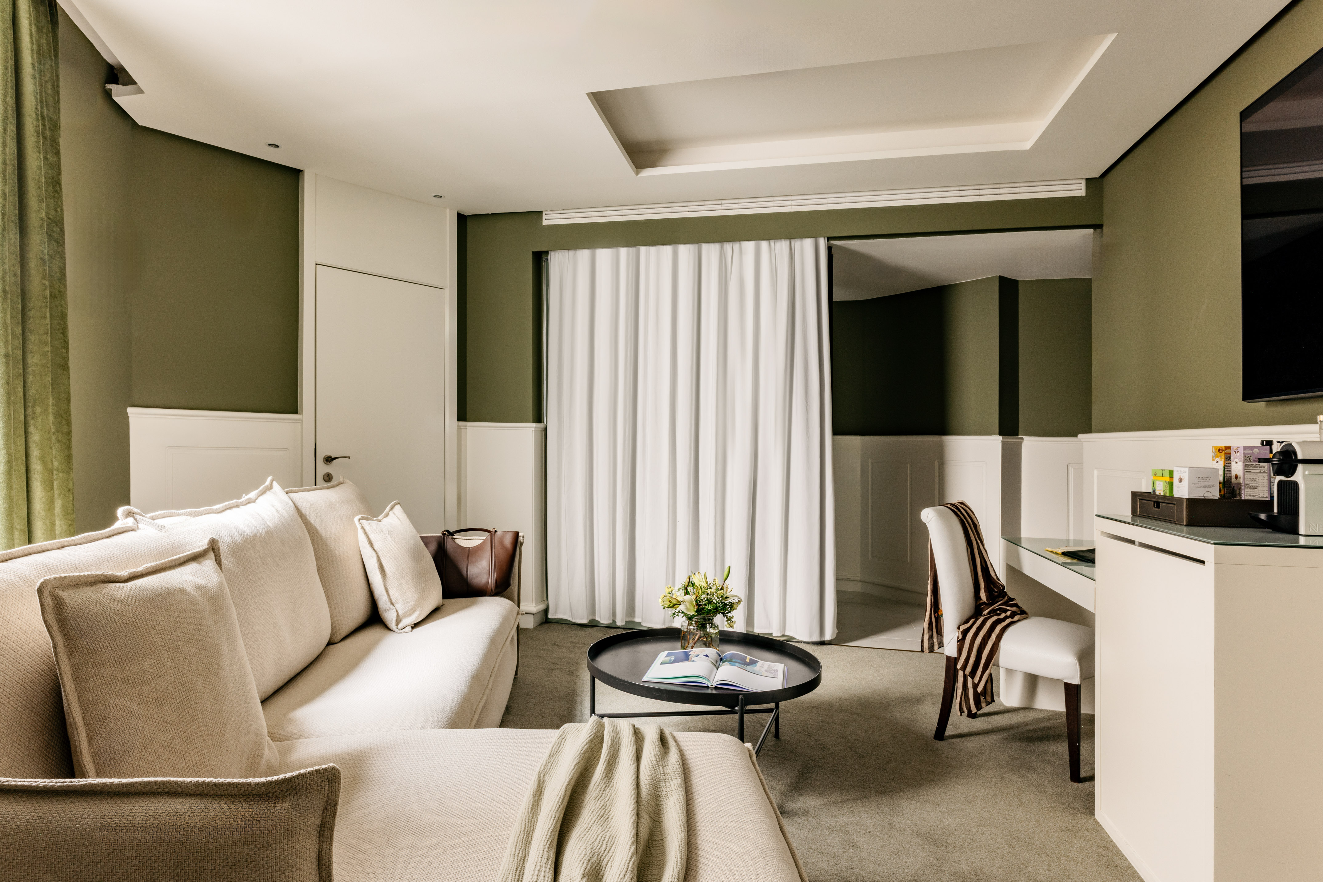 Suite de hotel elegante con sofá beige, mesa centro, paredes verde oliva.