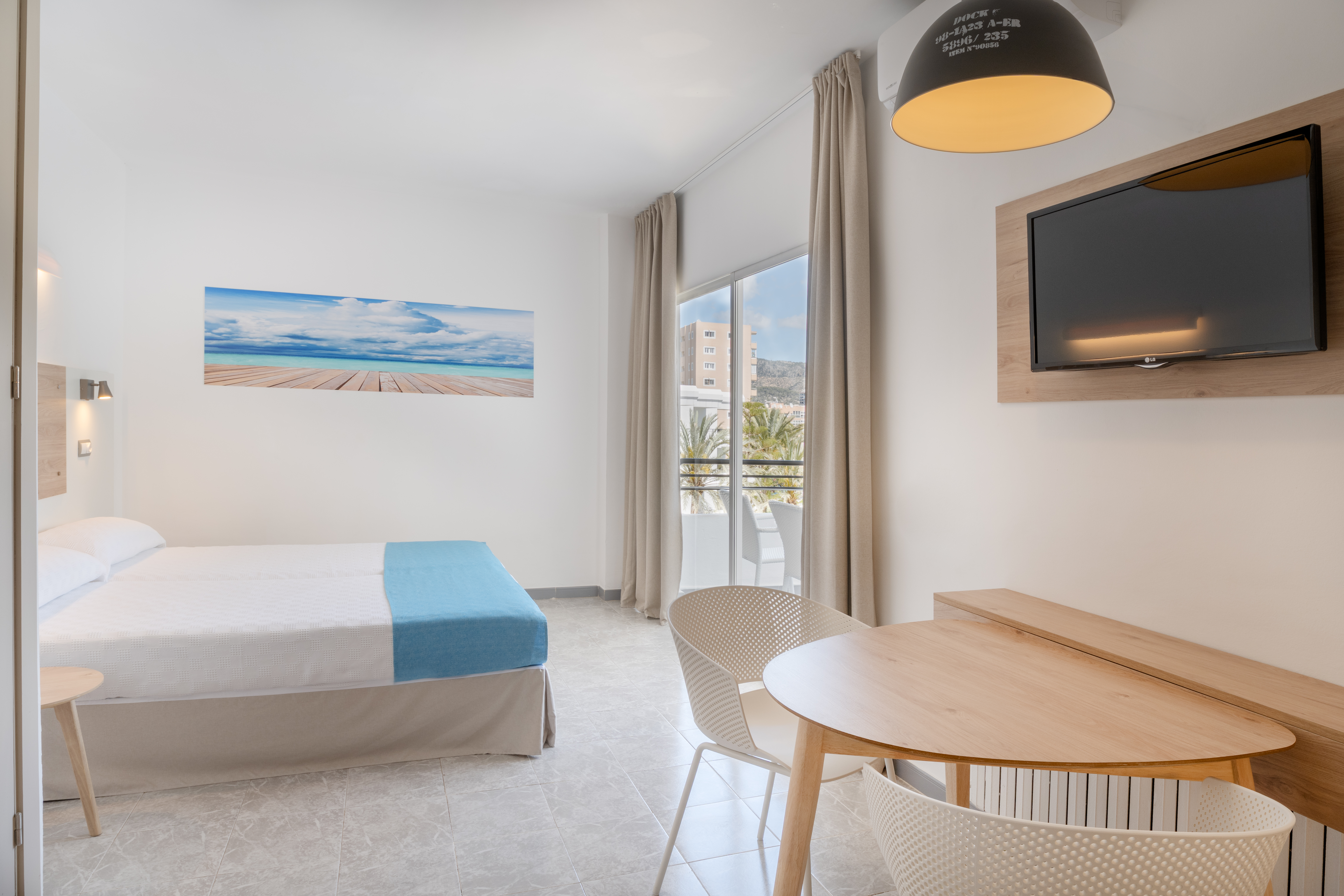 Modern hotel room with bed, TV, dining area, and balcony view of palm trees.