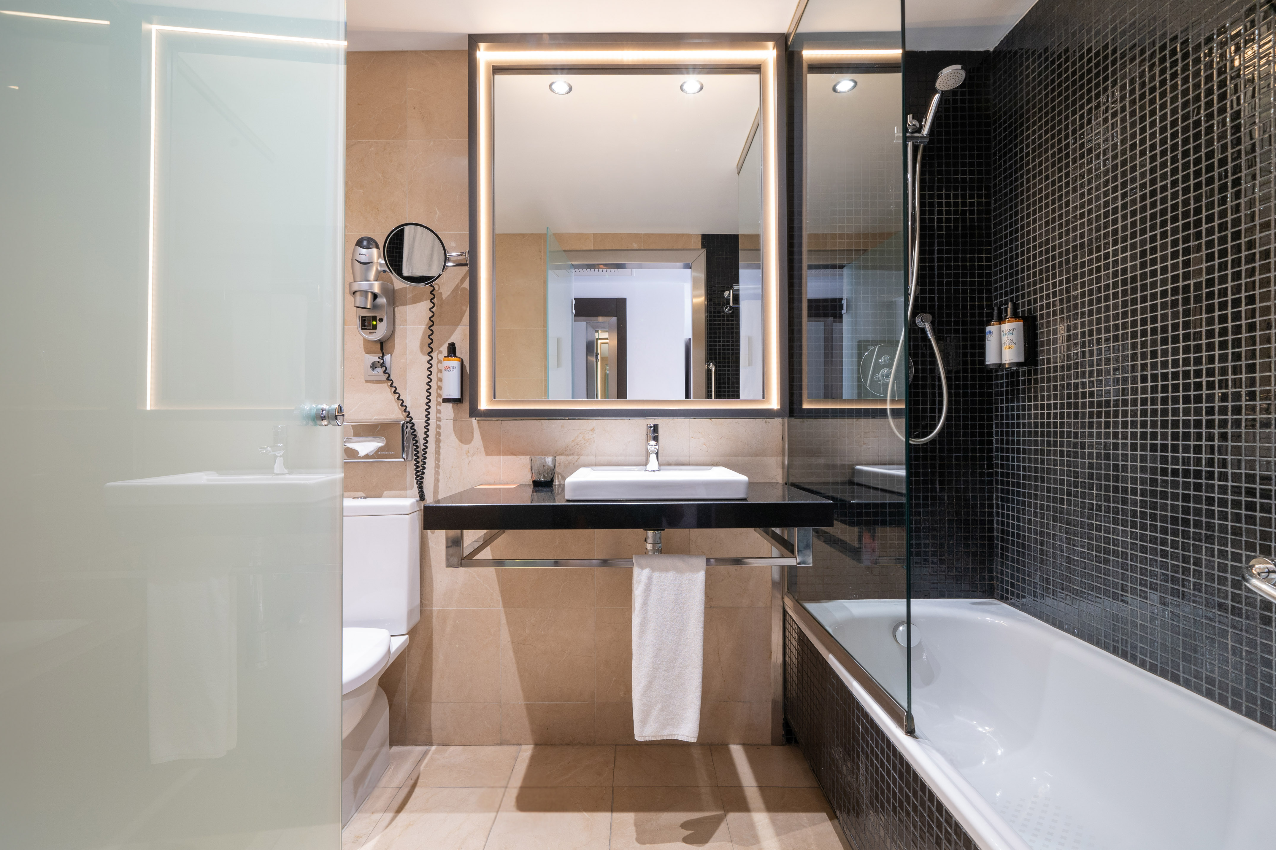 Modern bathroom with black mosaic shower, lighted mirror, and beige tiles.