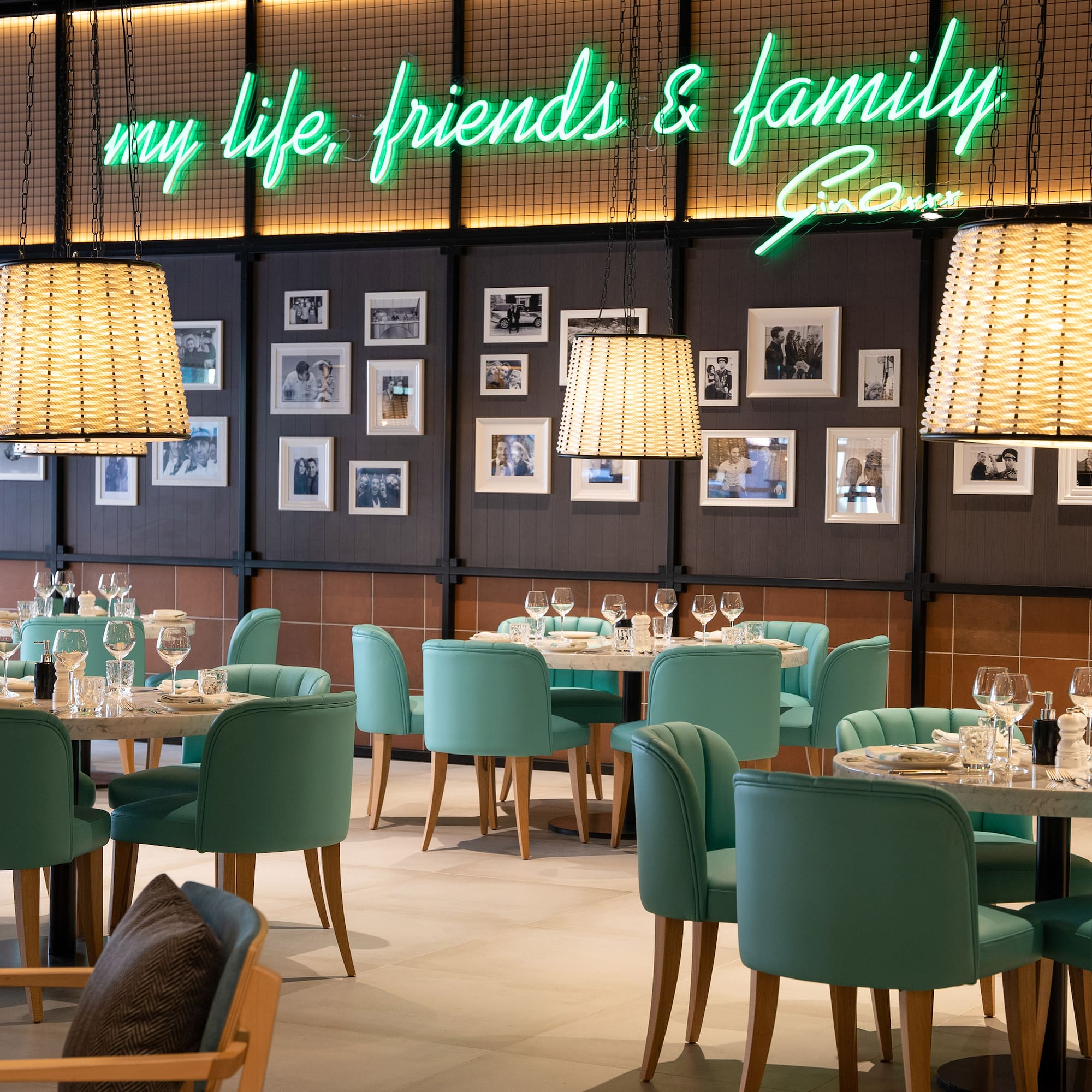 Restaurant interior with teal chairs, marble tables, framed photos, and green neon sign.