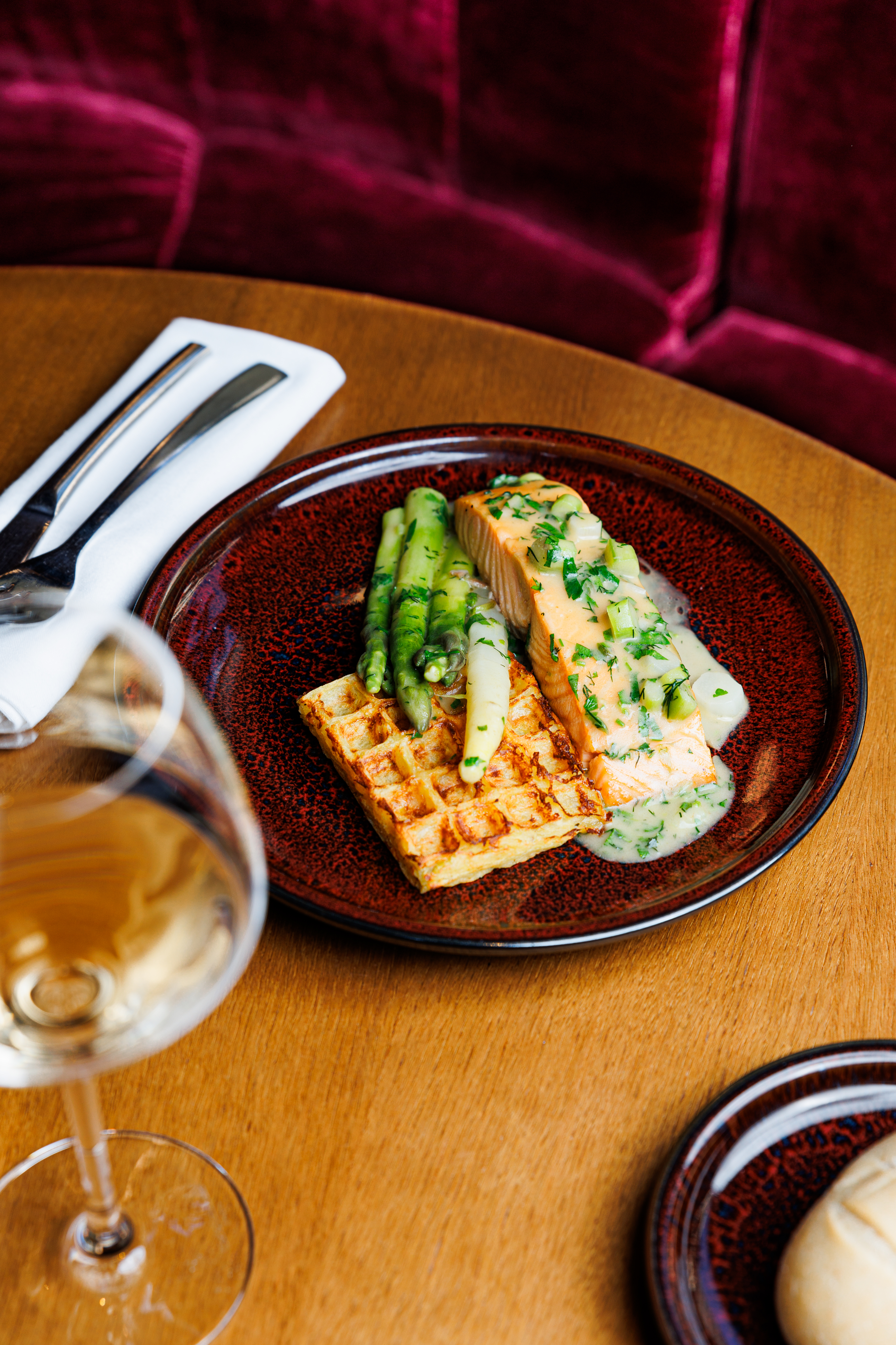 Salmon, green & white asparagus, waffle, and white wine on a dark red plate.