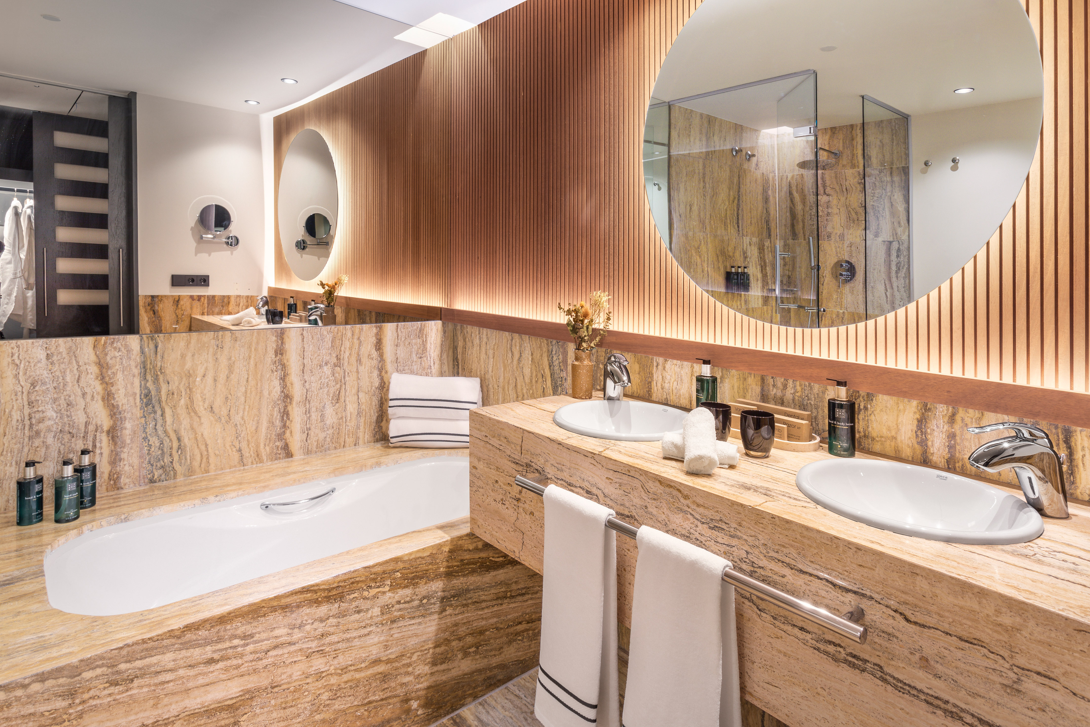 Modern luxury bathroom with stone bathtub, double vanity, and illuminated wood wall.
