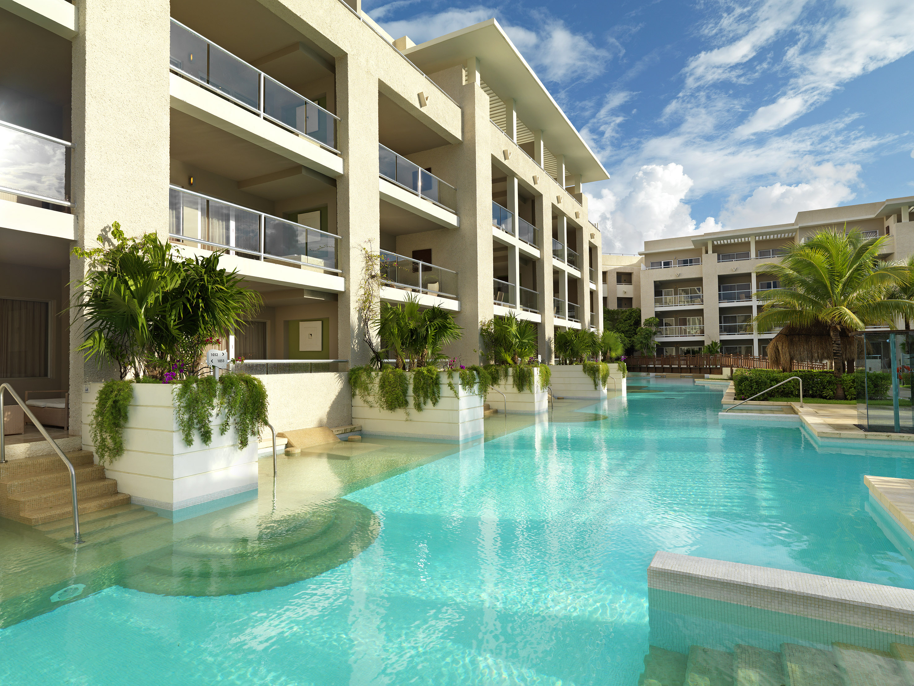 Modern hotel with turquoise swim-up pool, balconies, and tropical foliage.