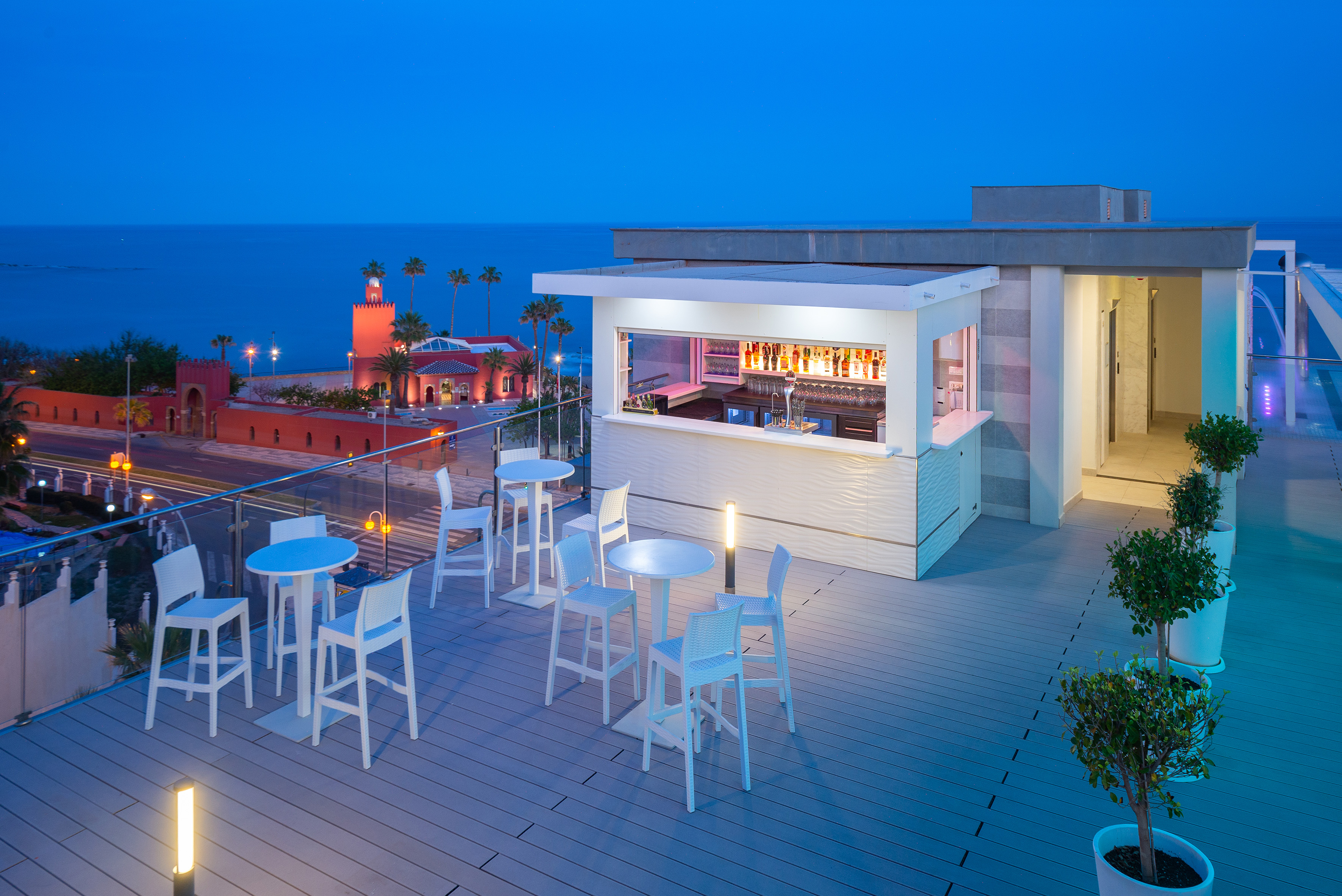 Coastal rooftop bar at night with ocean view, white tables, glowing counter.