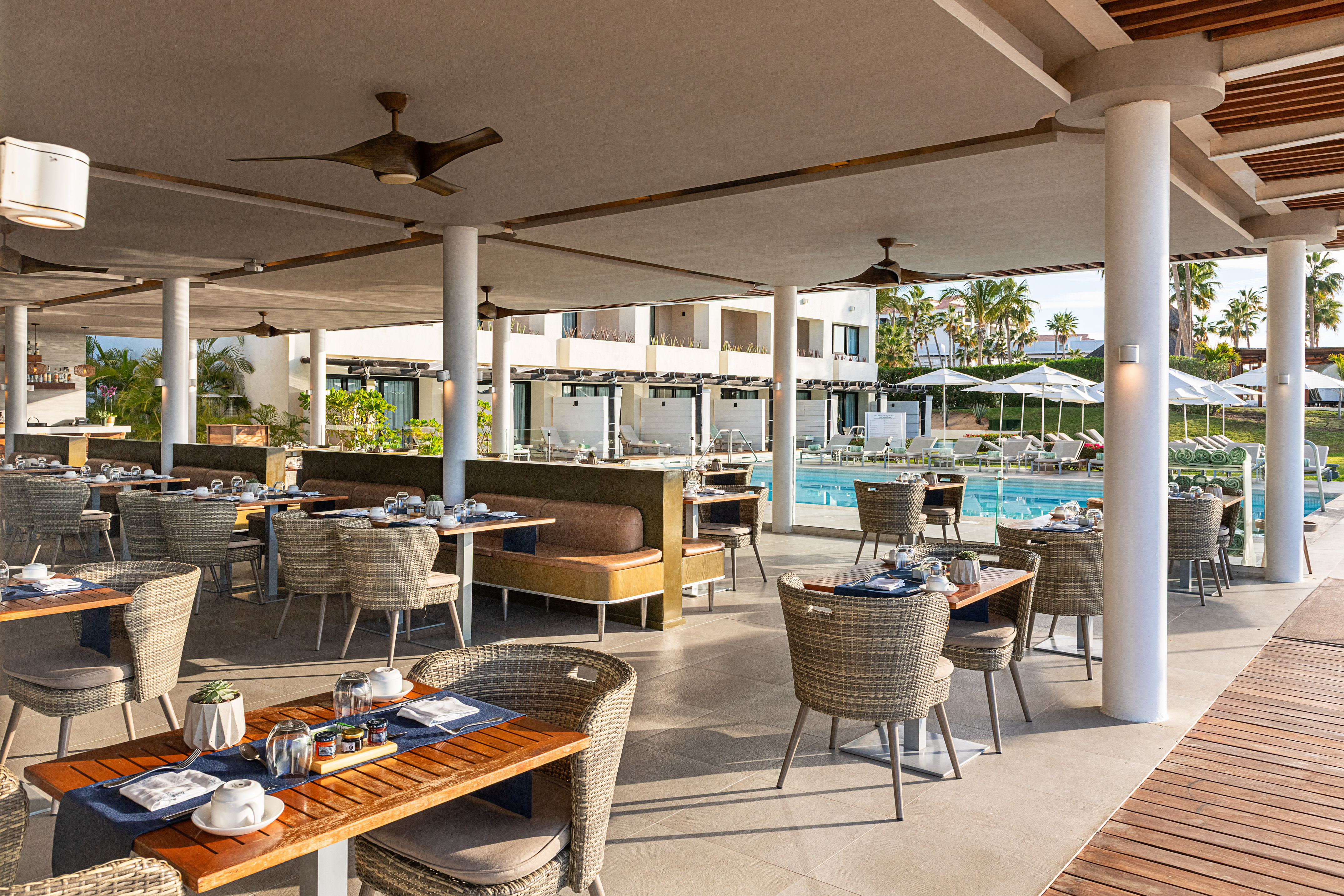 Outdoor resort restaurant with set tables, ceiling fans, pool, and palm trees.