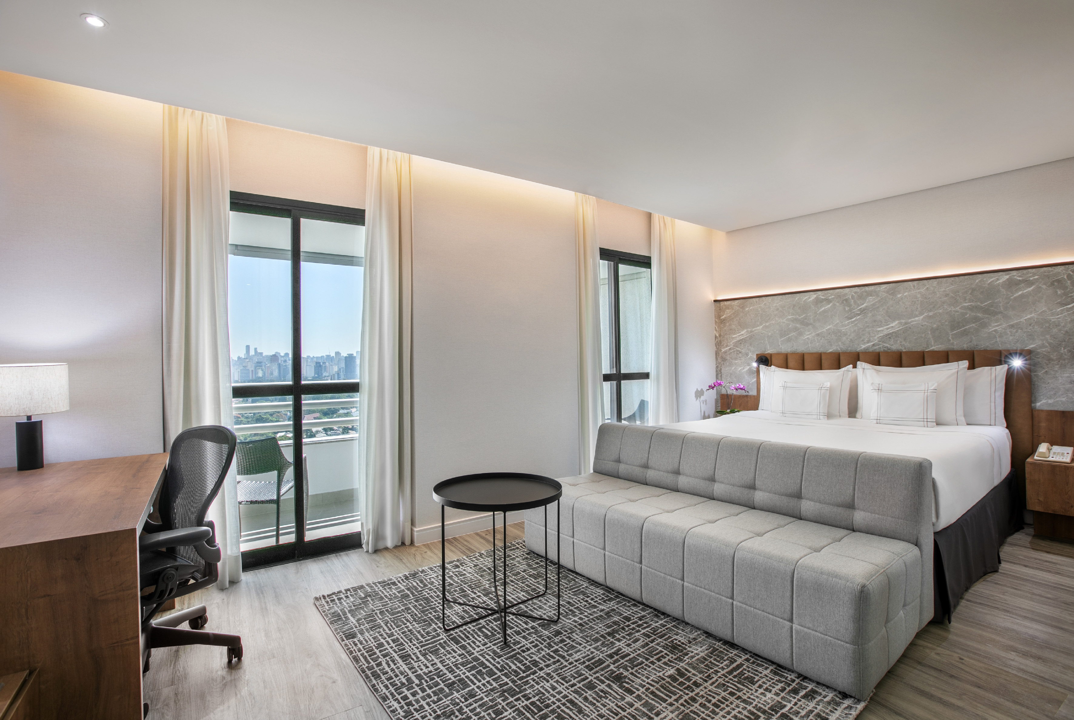 Modern hotel room with king bed, work desk, grey sofa, and city view balcony.