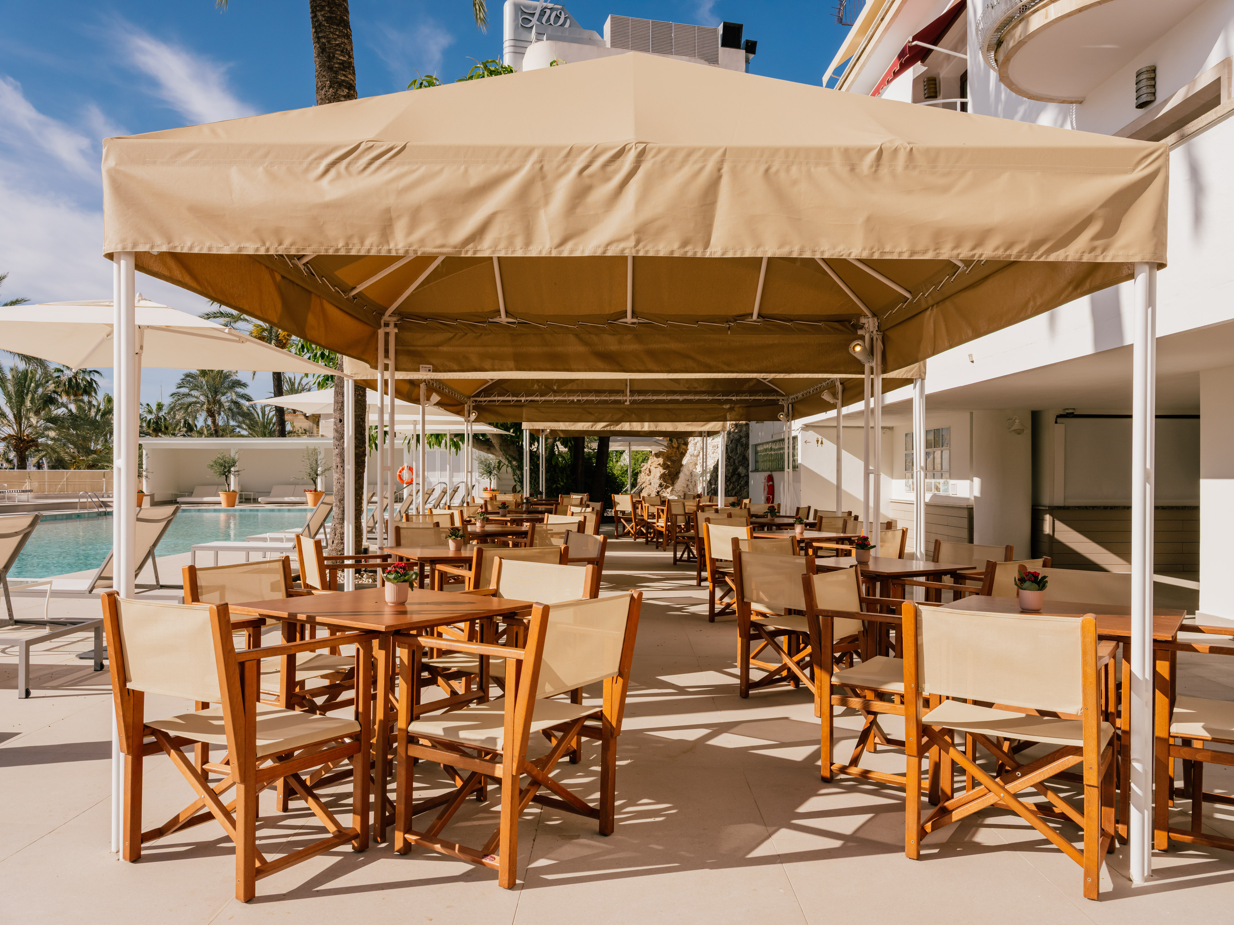 Shaded outdoor hotel restaurant patio with wooden tables and chairs, poolside view.