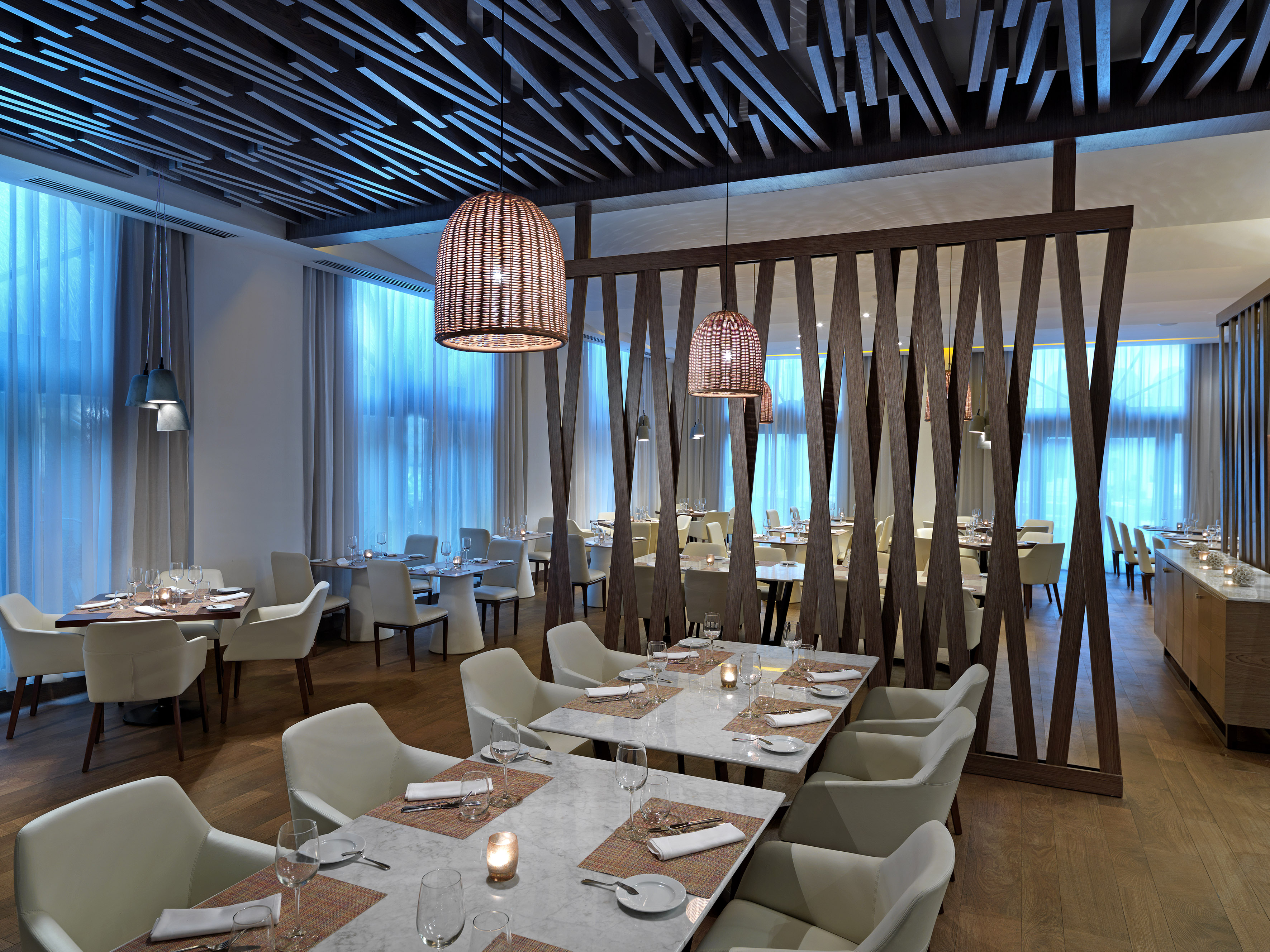 Modern restaurant dining with white chairs, woven pendants, and blue windows.