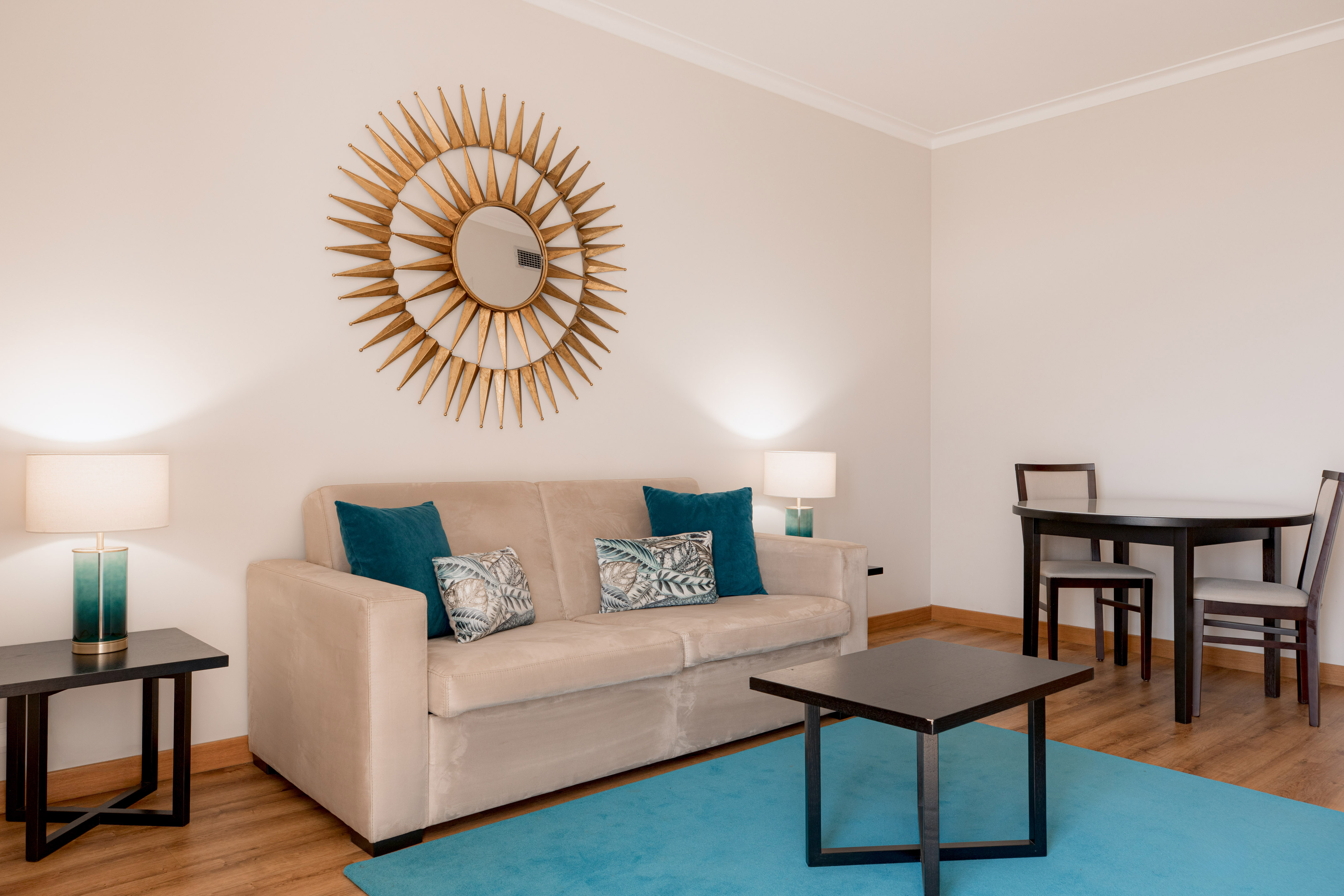 Cozy living room interior with beige sofa, gold sunburst mirror, and teal rug.