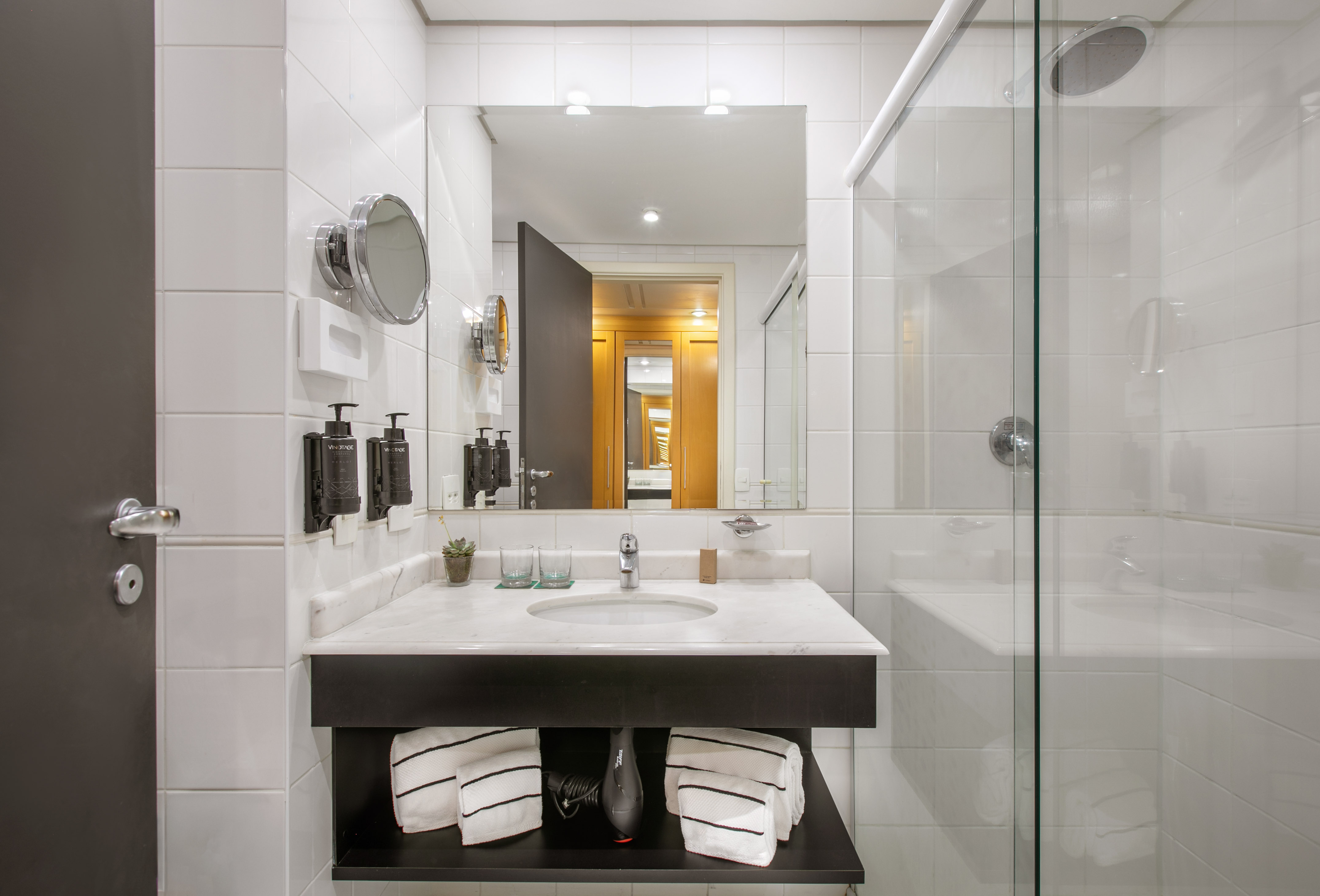 Modern hotel bathroom with white tiles, marble vanity, sink, and glass shower.