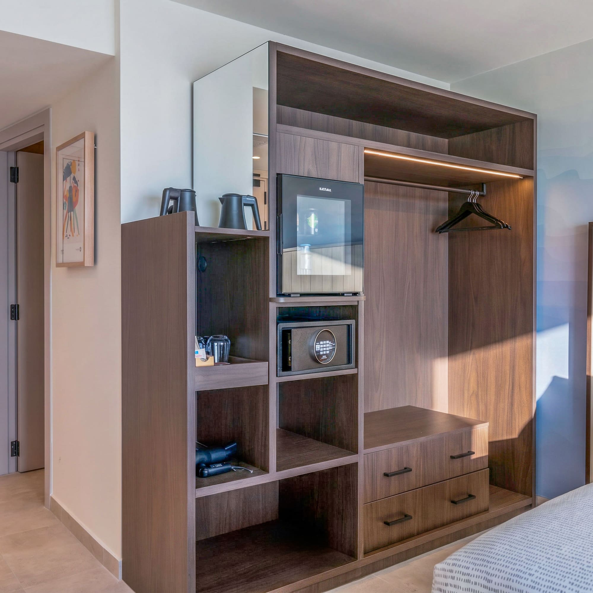 Modern hotel room with built-in dark wood wardrobe, mini-fridge, and safe.