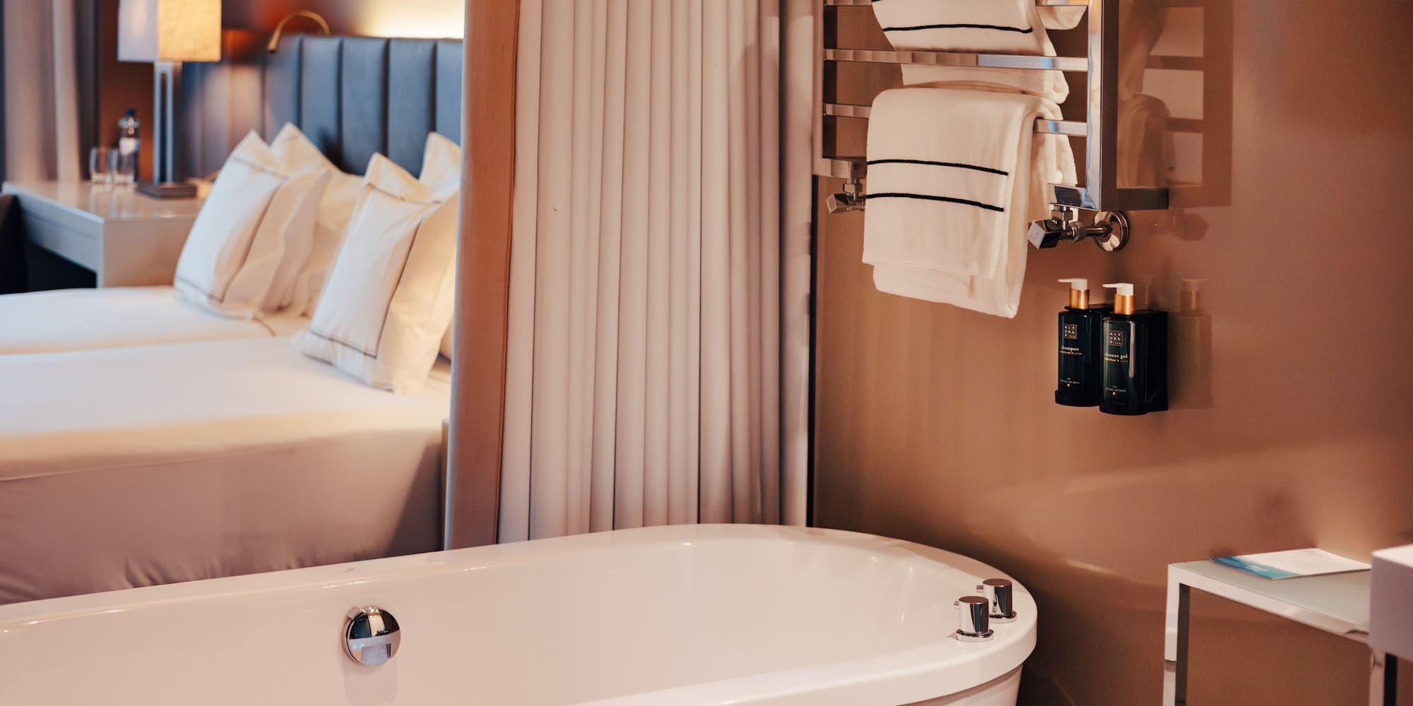 Modern hotel suite: sleek white bathtub, heated towel rack, and cozy bed.