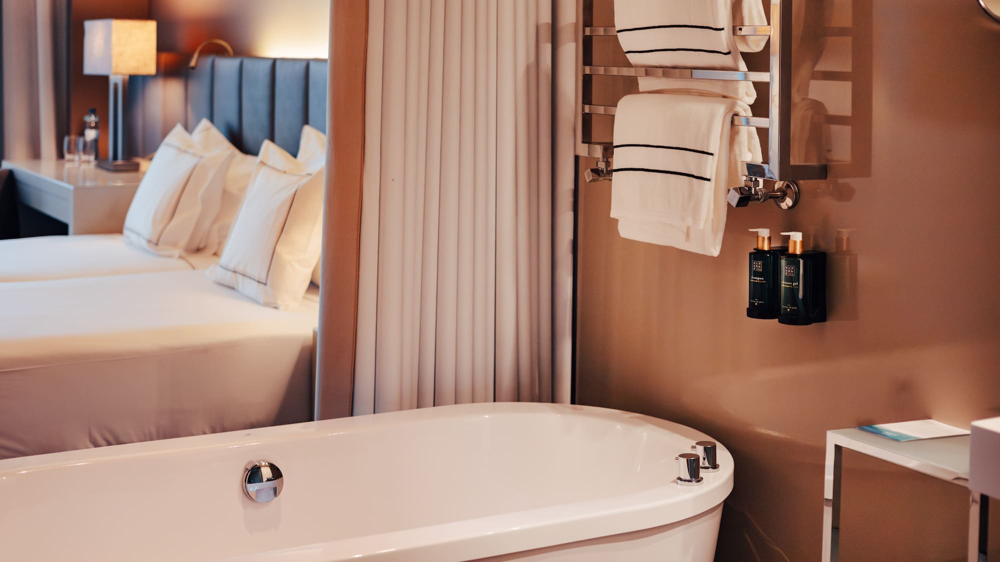 Modern hotel suite: sleek white bathtub, heated towel rack, and cozy bed.