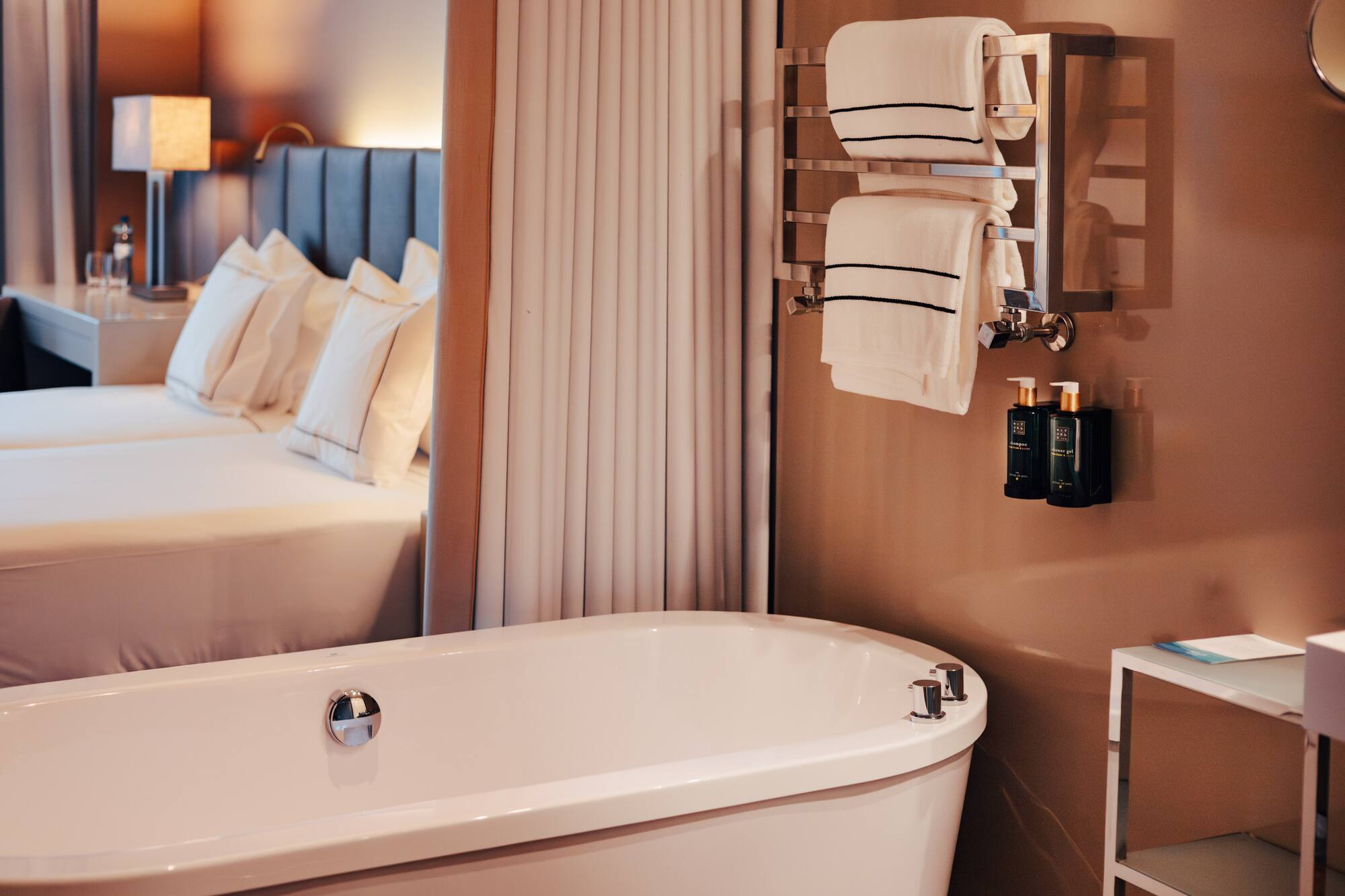 Modern hotel suite: sleek white bathtub, heated towel rack, and cozy bed.