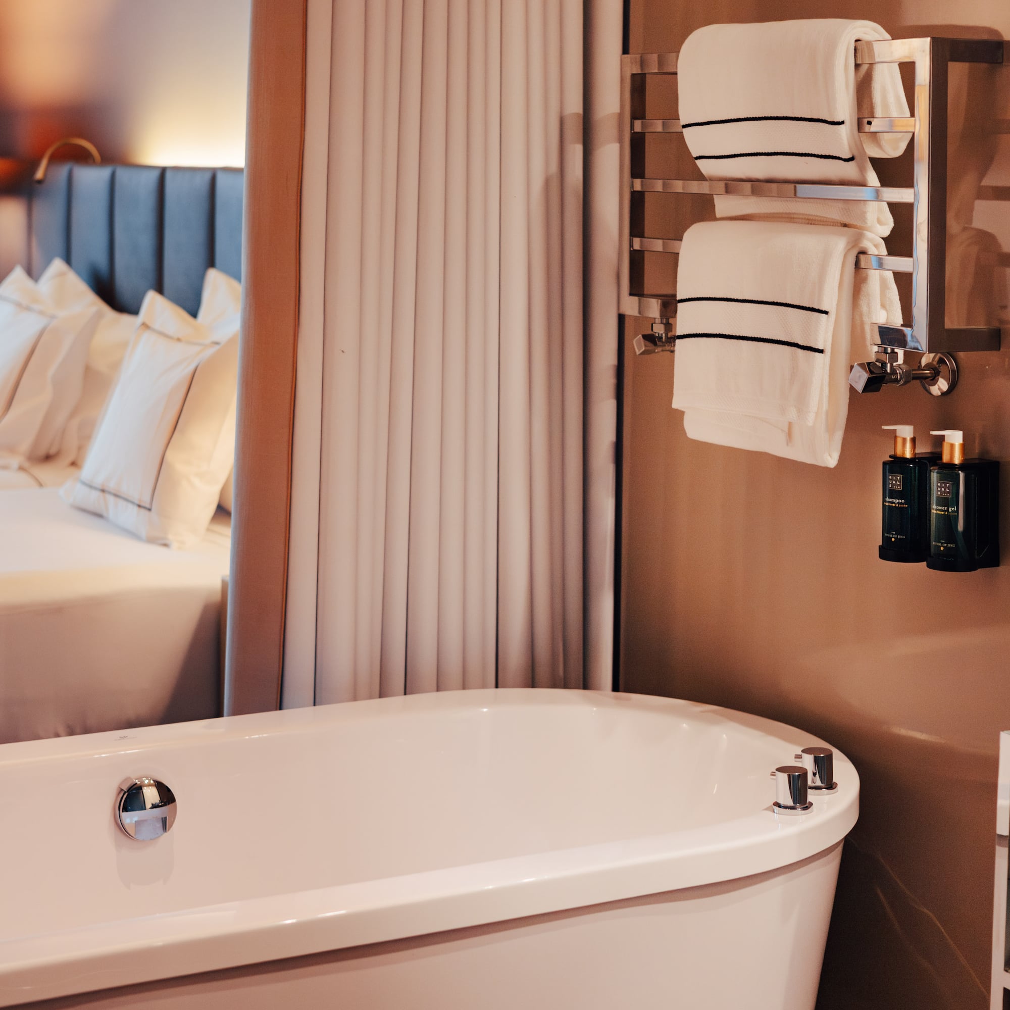 Modern hotel suite: sleek white bathtub, heated towel rack, and cozy bed.