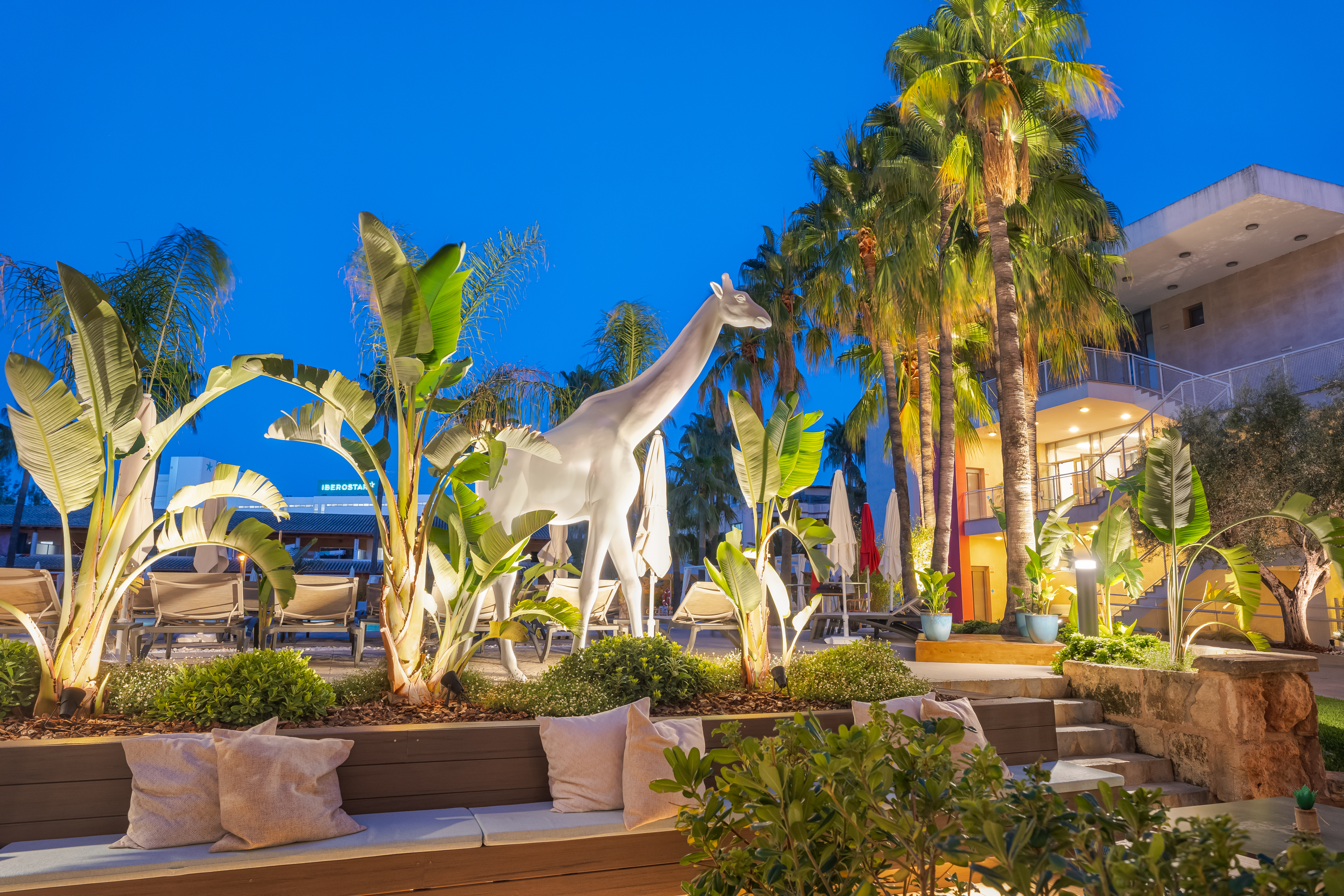 Night tropical resort: palm trees, white giraffe statue, outdoor lounge.