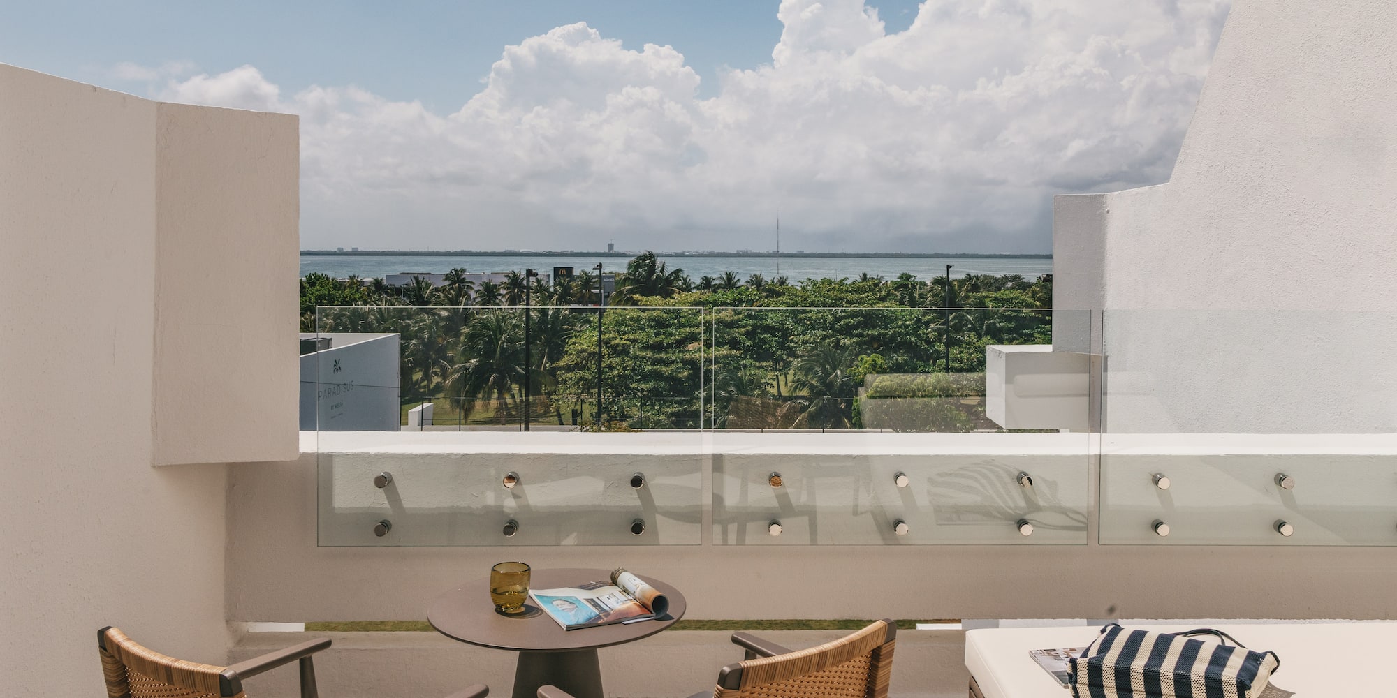 Modern hotel balcony with ocean view, tropical trees, and outdoor seating.