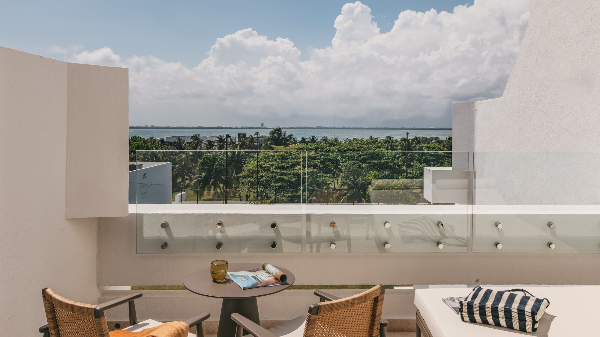 Modern hotel balcony with ocean view, tropical trees, and outdoor seating.