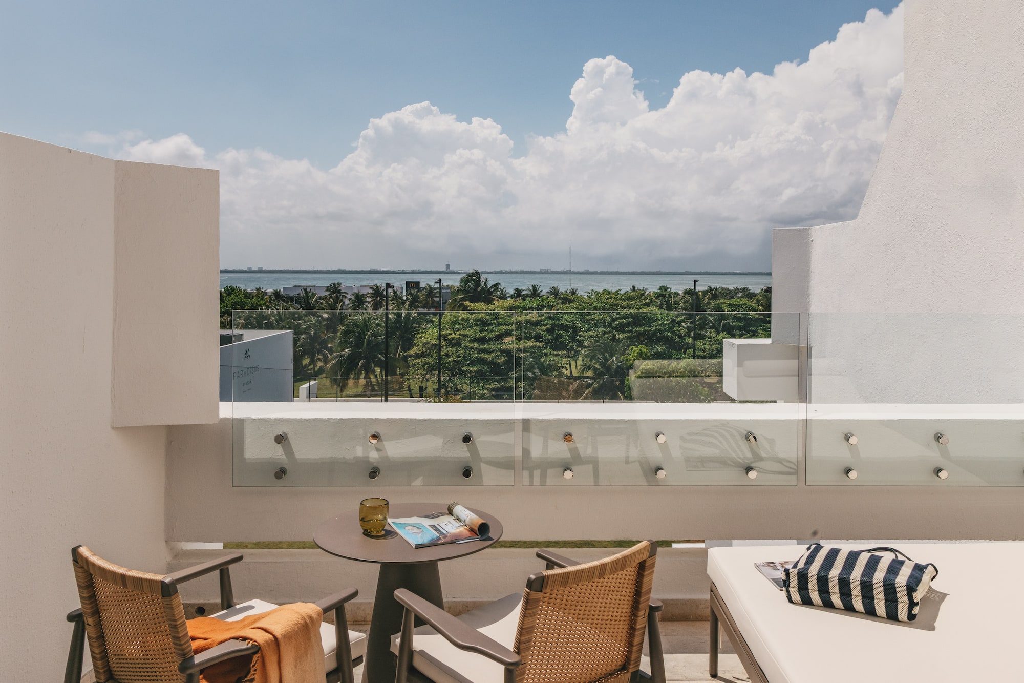 Modern hotel balcony with ocean view, tropical trees, and outdoor seating.