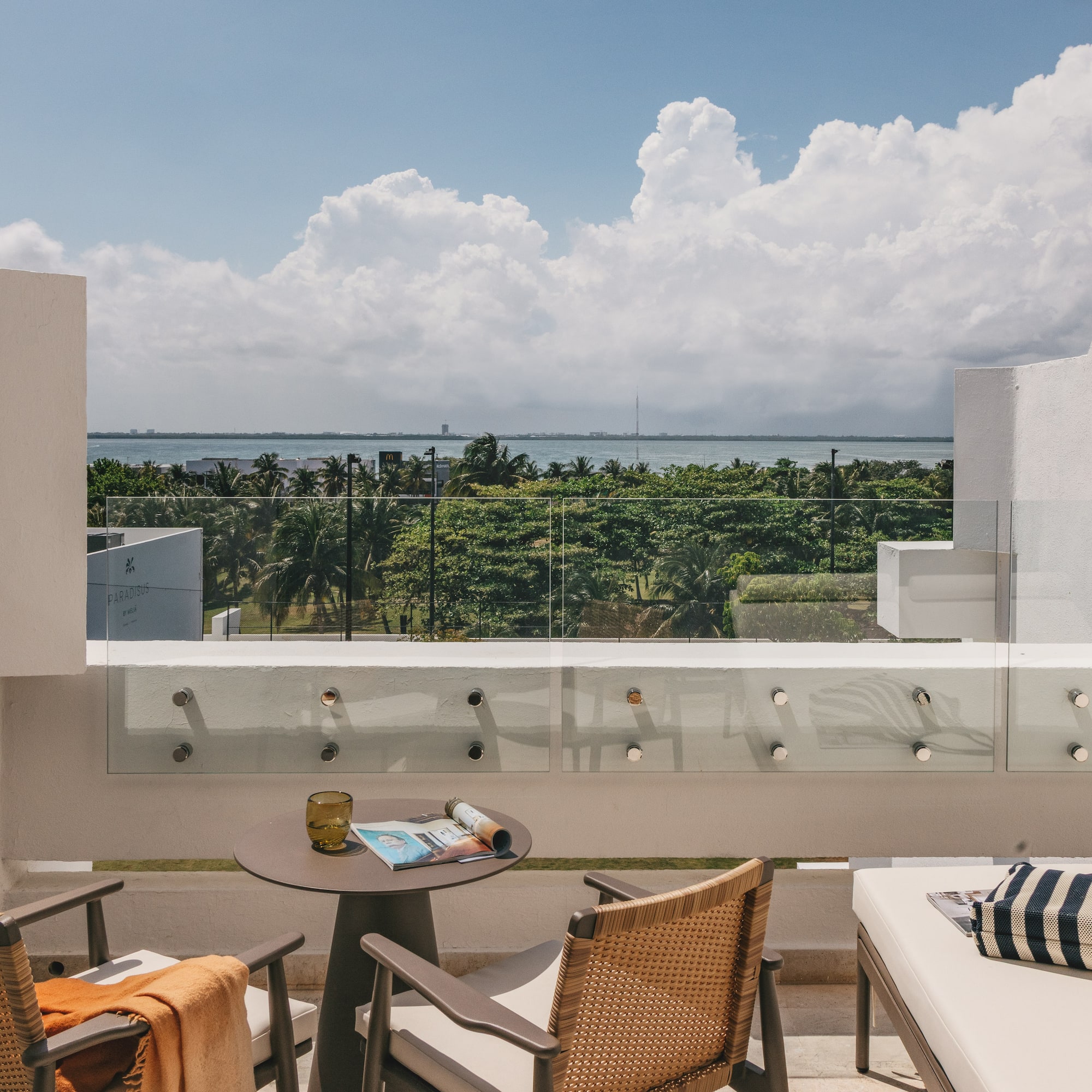 Modern hotel balcony with ocean view, outdoor seating, and palm trees.
