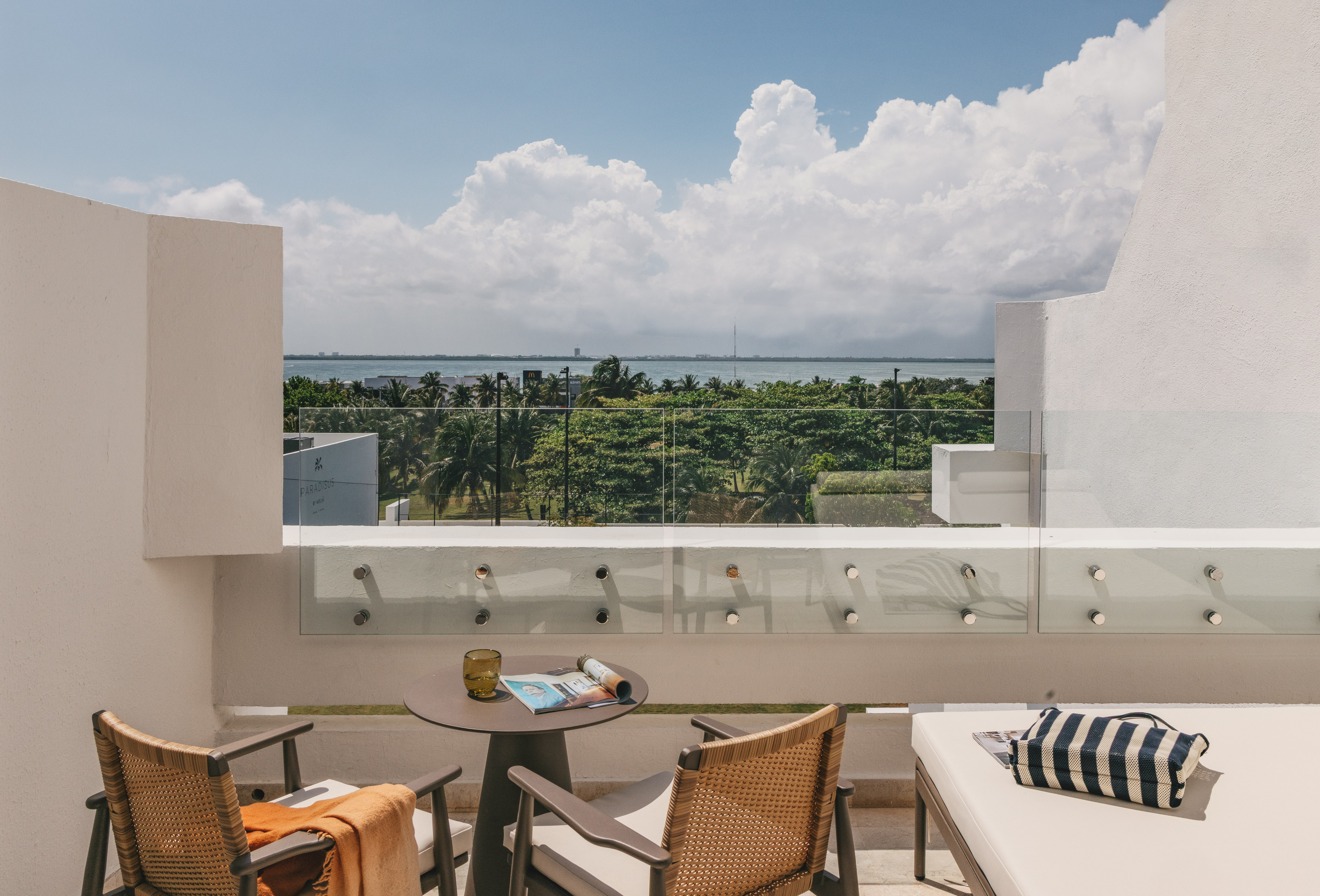 Modern hotel balcony with ocean view, tropical trees, and outdoor seating.