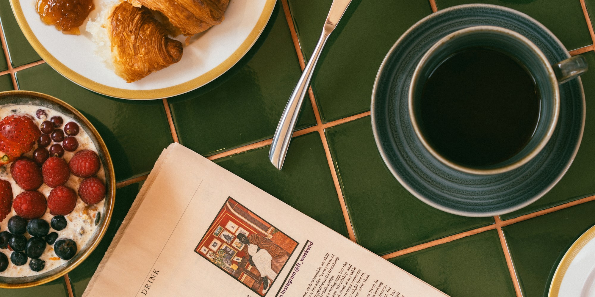 Breakfast with croissants, coffee, fruit, berries, and newspaper on green tiles.