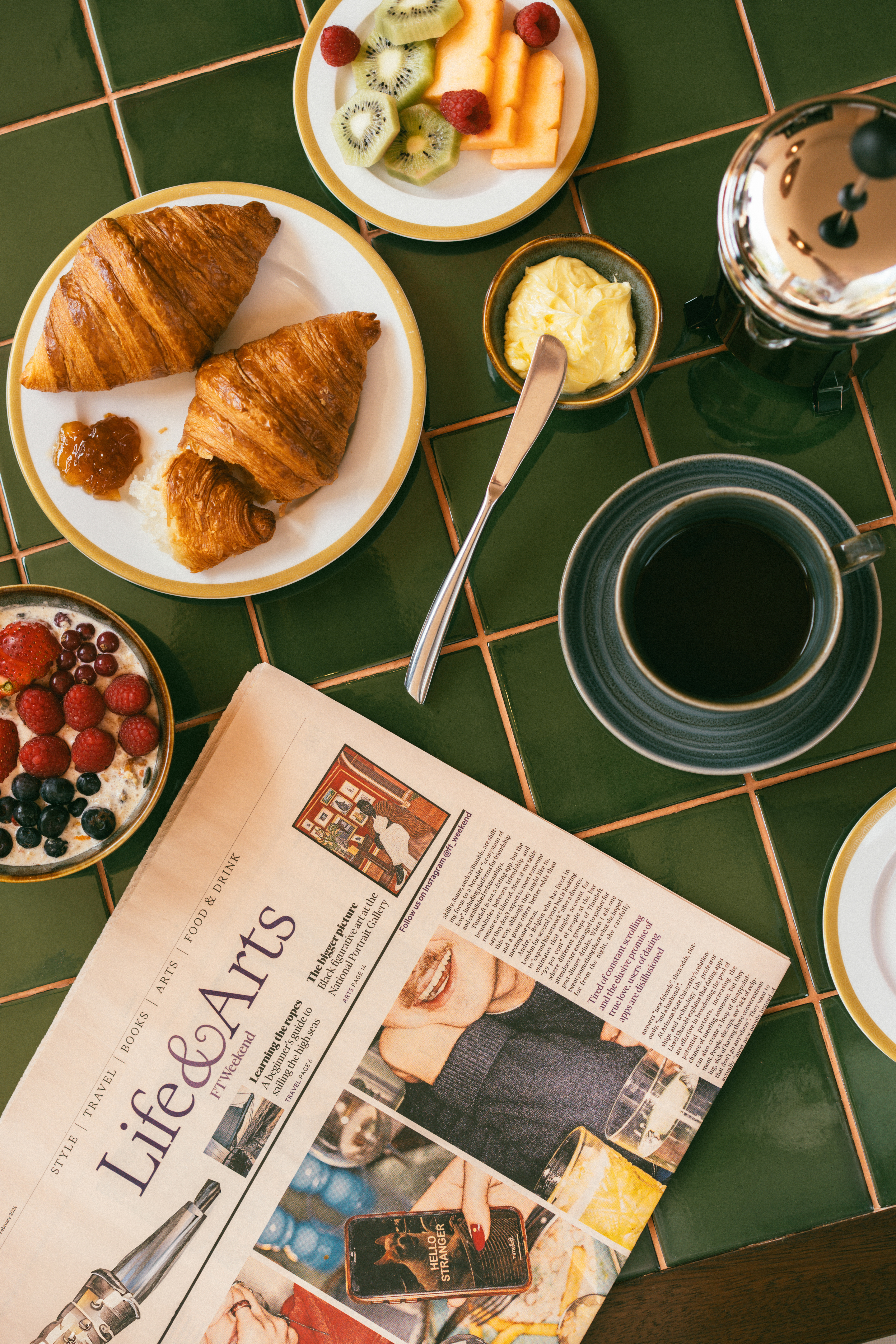Breakfast with croissants, coffee, fruit, berries, and newspaper on green tiles.