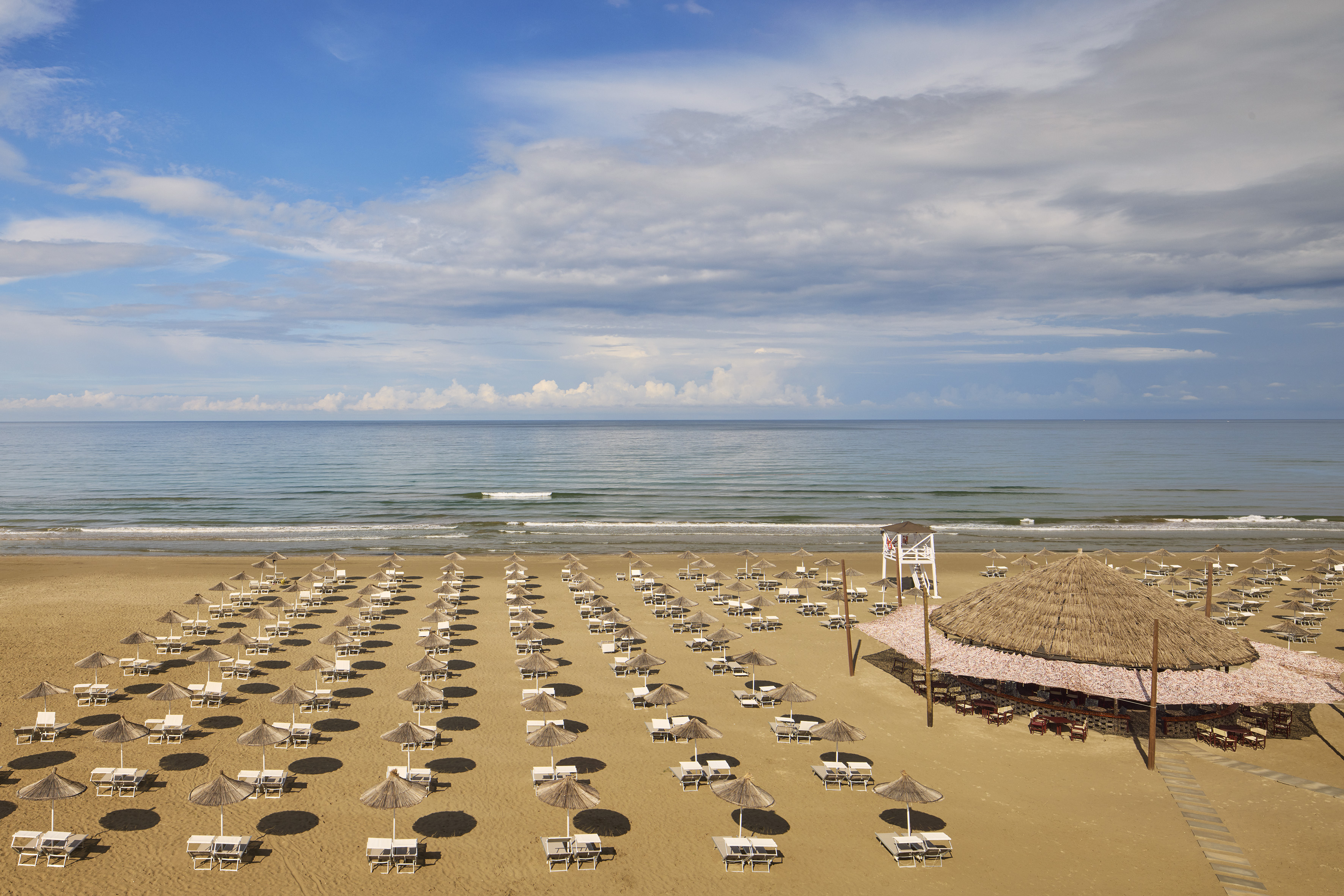 Beach resort: rows of straw umbrellas, loungers, blue ocean, thatched bar.