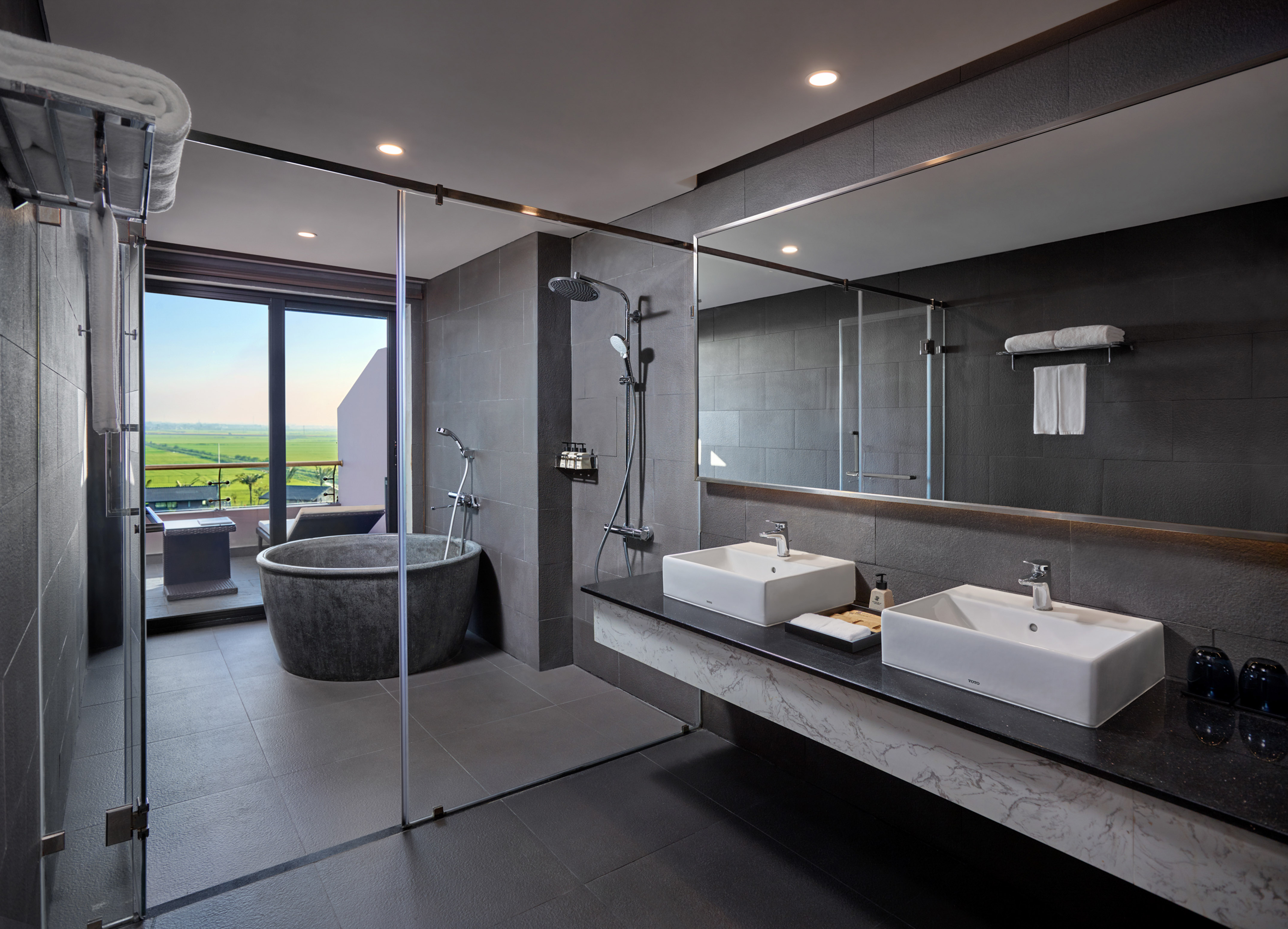Modern hotel bathroom with stone tub, double sinks, glass shower, and scenic view.